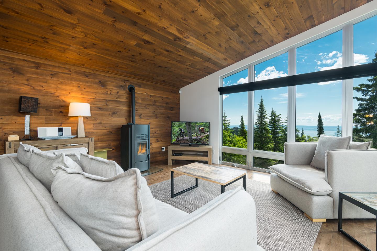 Salon moderne avec grandes fenêtres, vue sur forêt et lac, canapés blancs, plafond en bois.