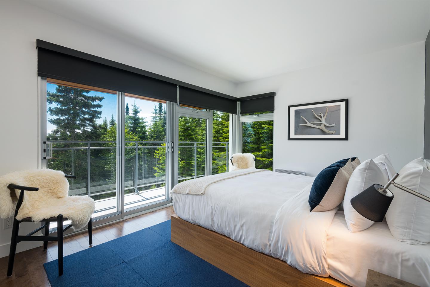 Chambre moderne, lit blanc, grandes fenêtres avec vue sur forêt.