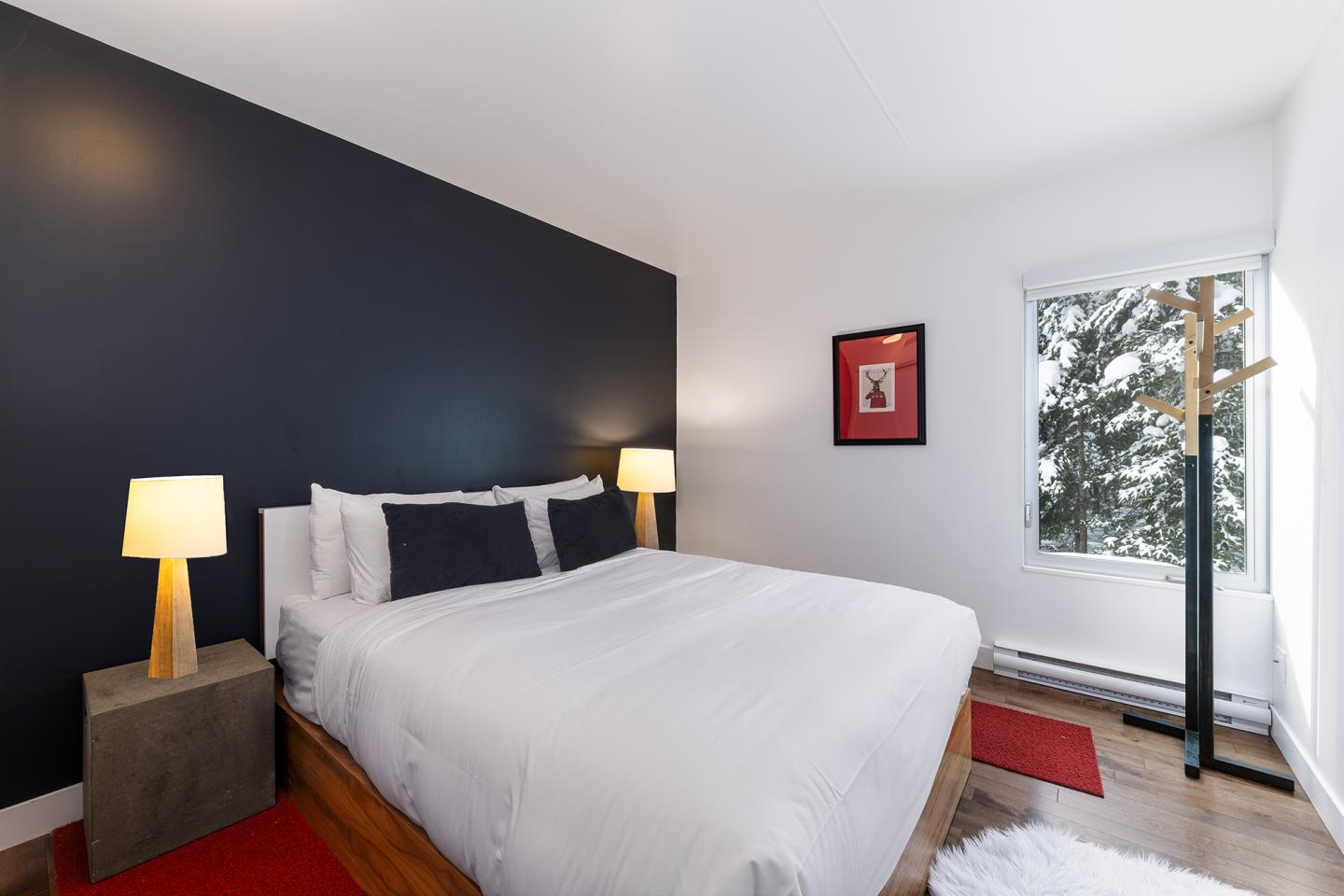 Chambre moderne avec lit double, murs sombres, lampes allumées et fenêtre sur paysage enneigé.