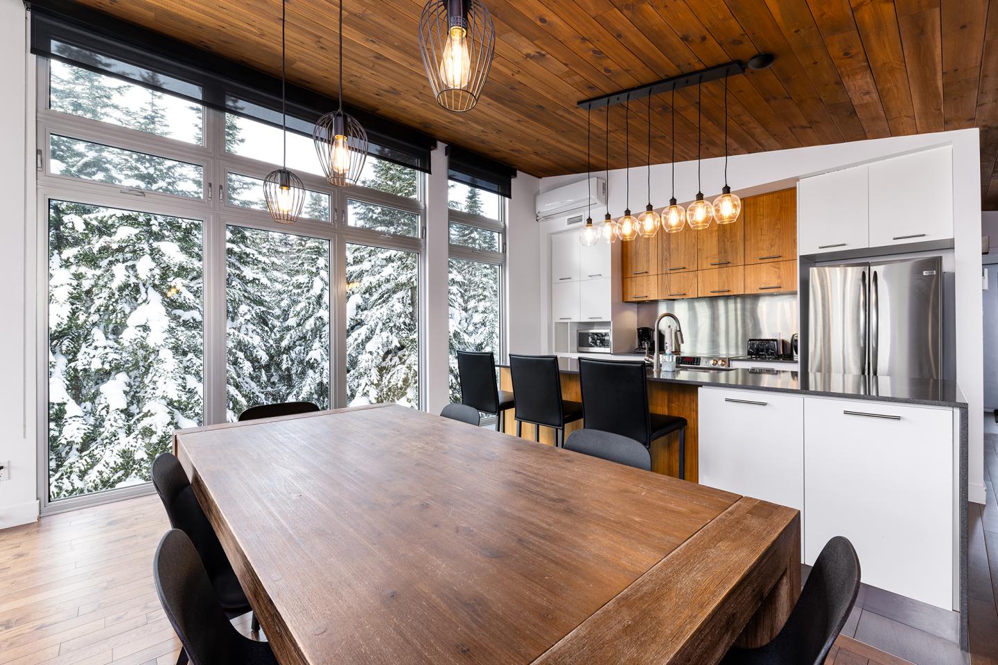 Cuisine moderne avec table en bois, grandes fenêtres, vue sur arbres enneigés.