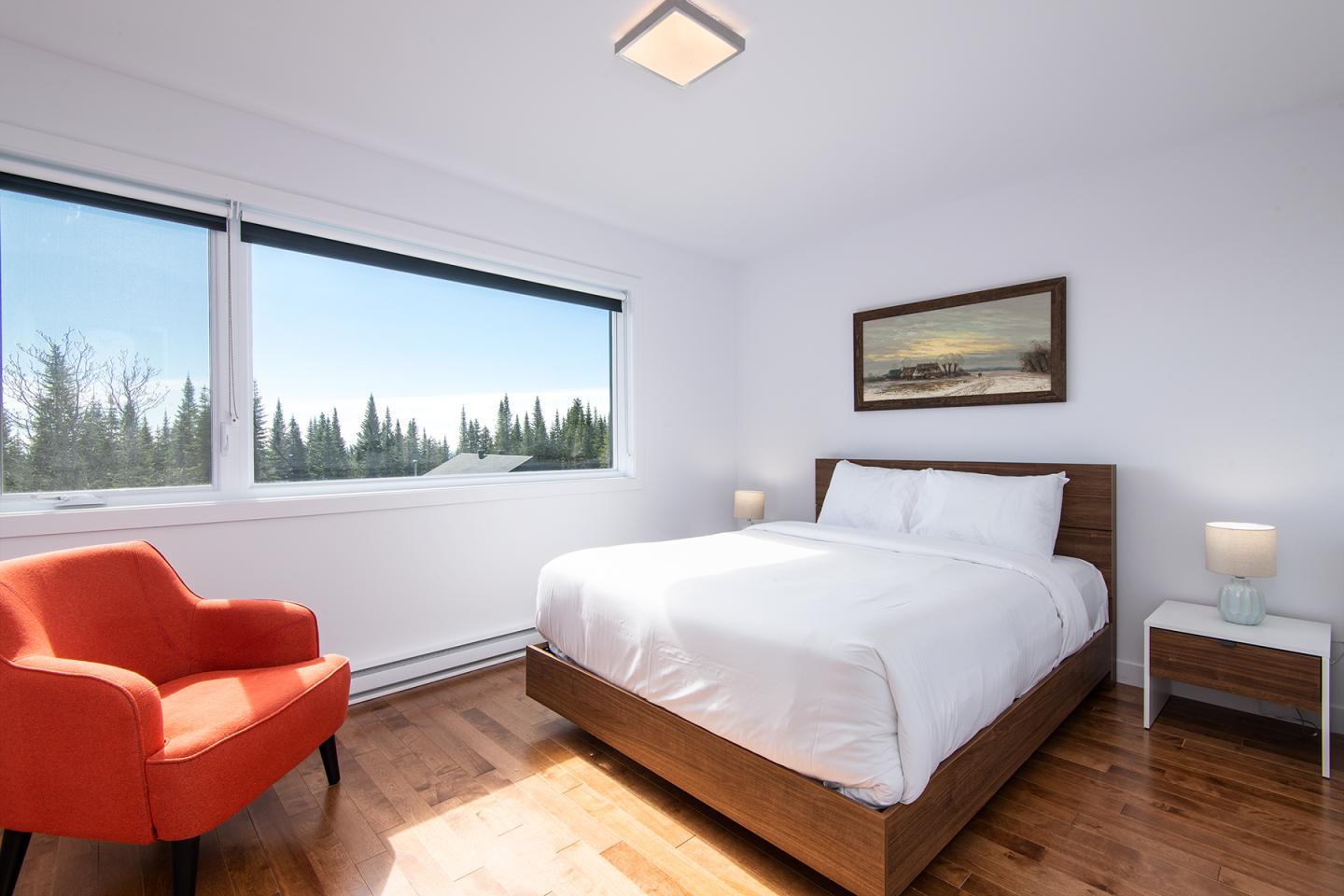 Chambre lumineuse, lit blanc, fauteuil orange, vue sur arbres.