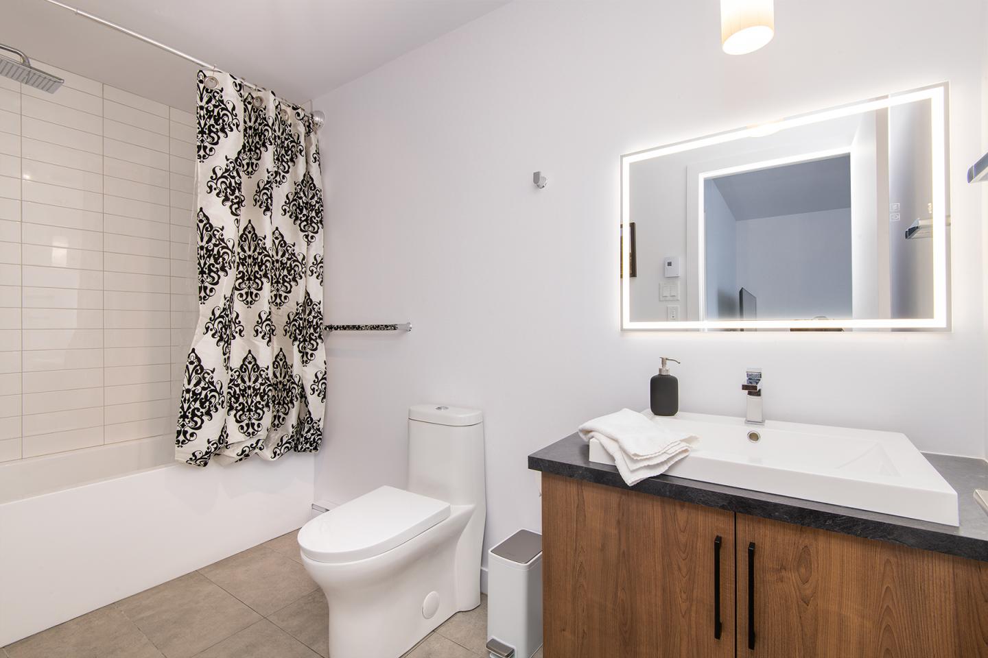 Salle de bain moderne avec lavabo, miroir éclairé et rideau de douche à motifs.