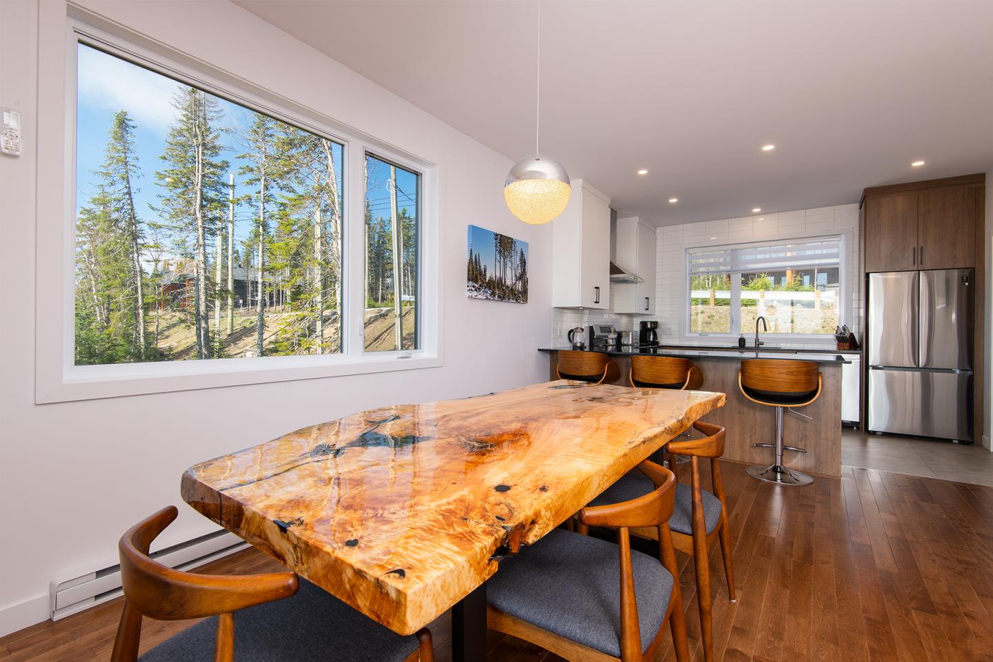 Cuisine moderne avec table en bois, chaises, et vue sur la forêt par la fenêtre.