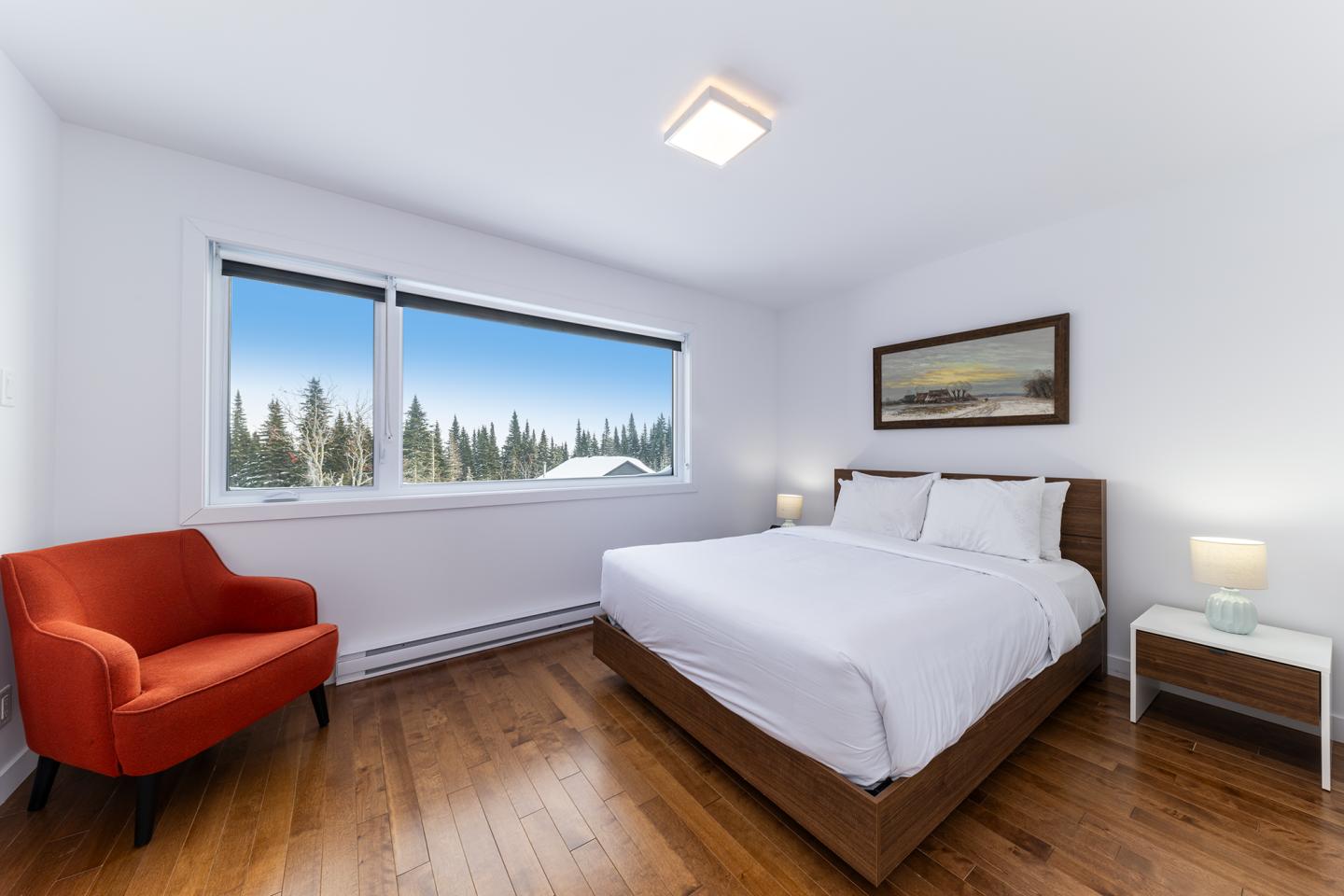 Chambre lumineuse avec lit double, fauteuil rouge, tableau et fenêtre avec vue sur arbres.