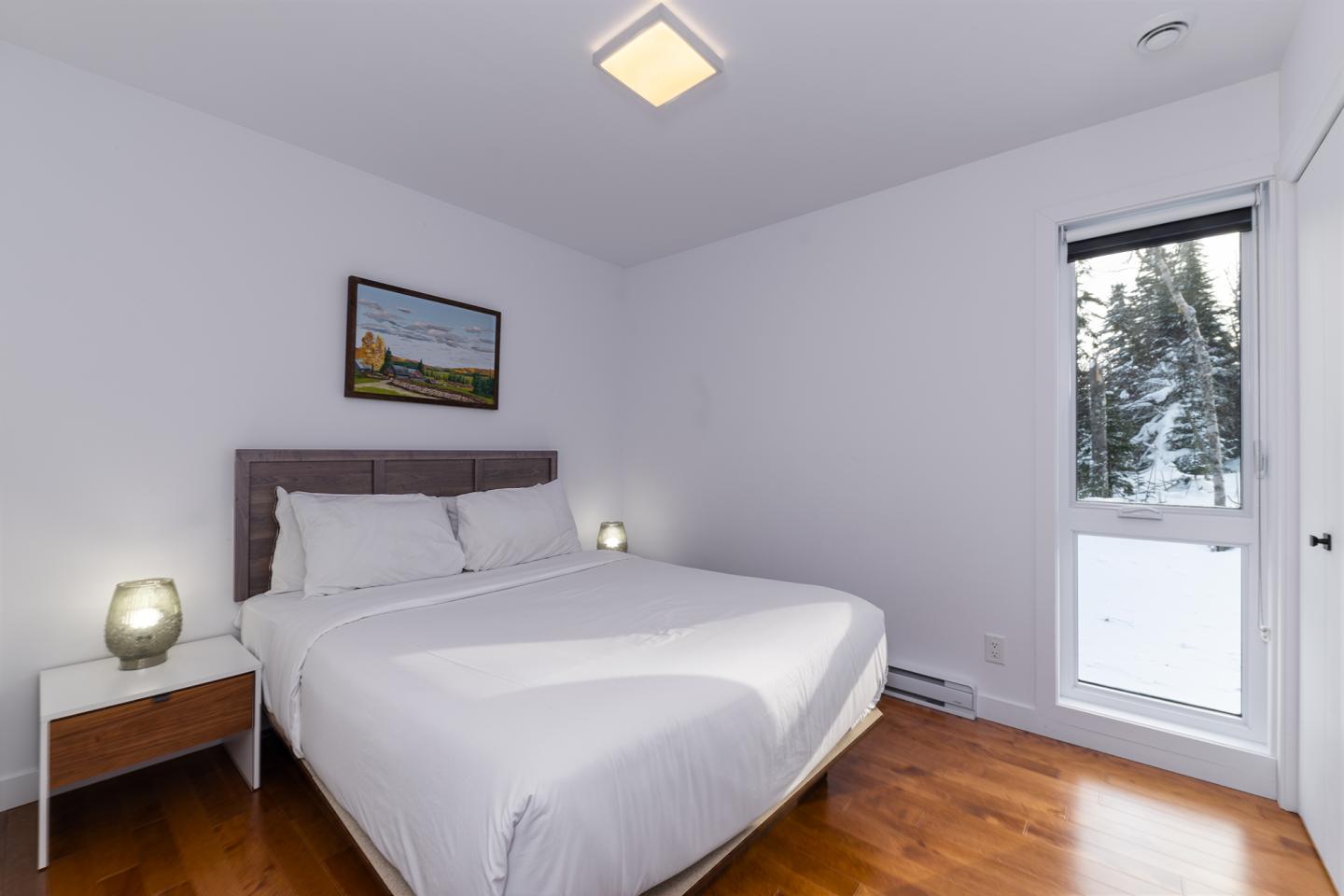 Chambre minimaliste, lit double, murs blancs, parquet, fenêtre donnant sur un paysage enneigé.