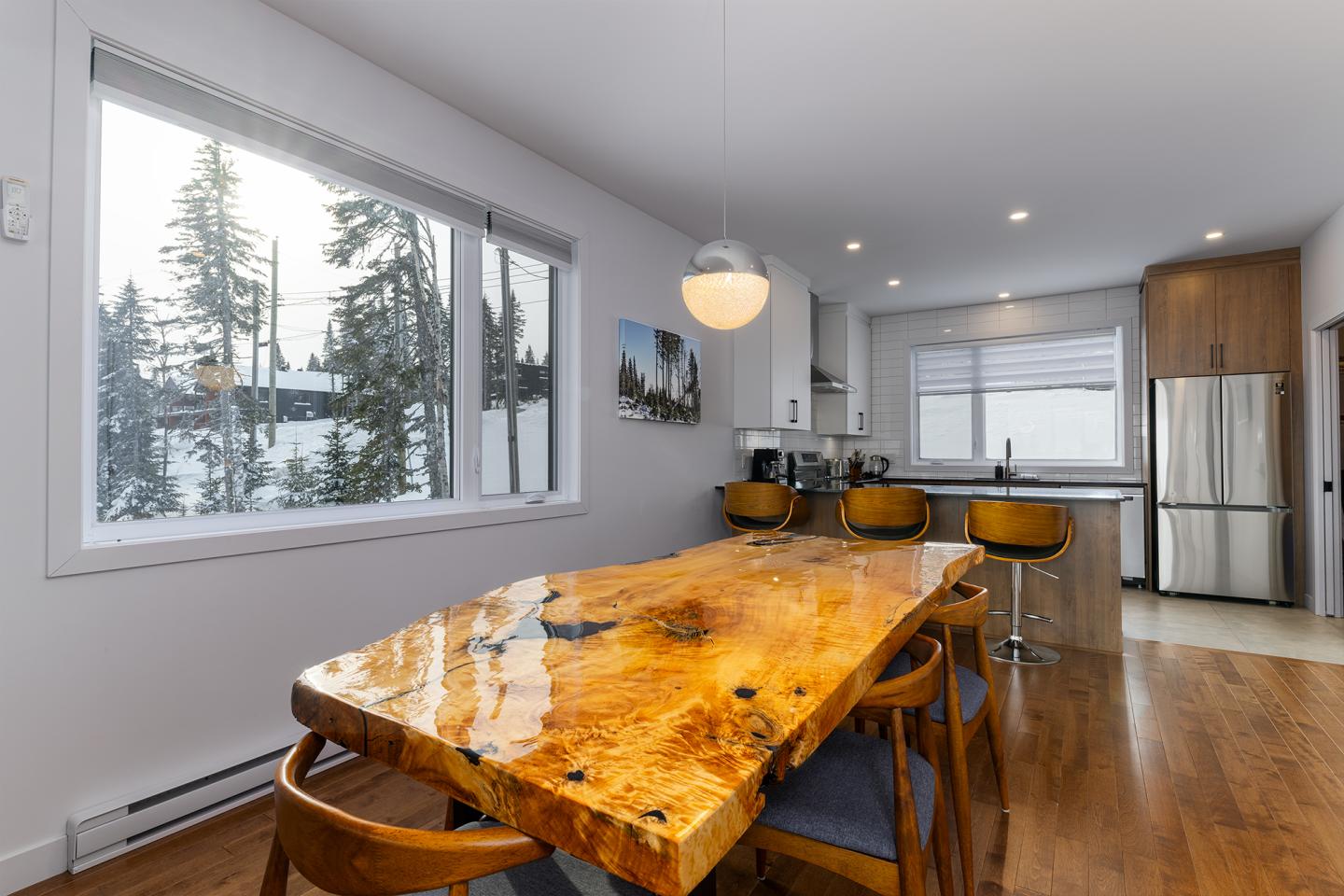 Salle à manger moderne avec table en bois, chaises et cuisine ouverte, vue sur des arbres enneigés.