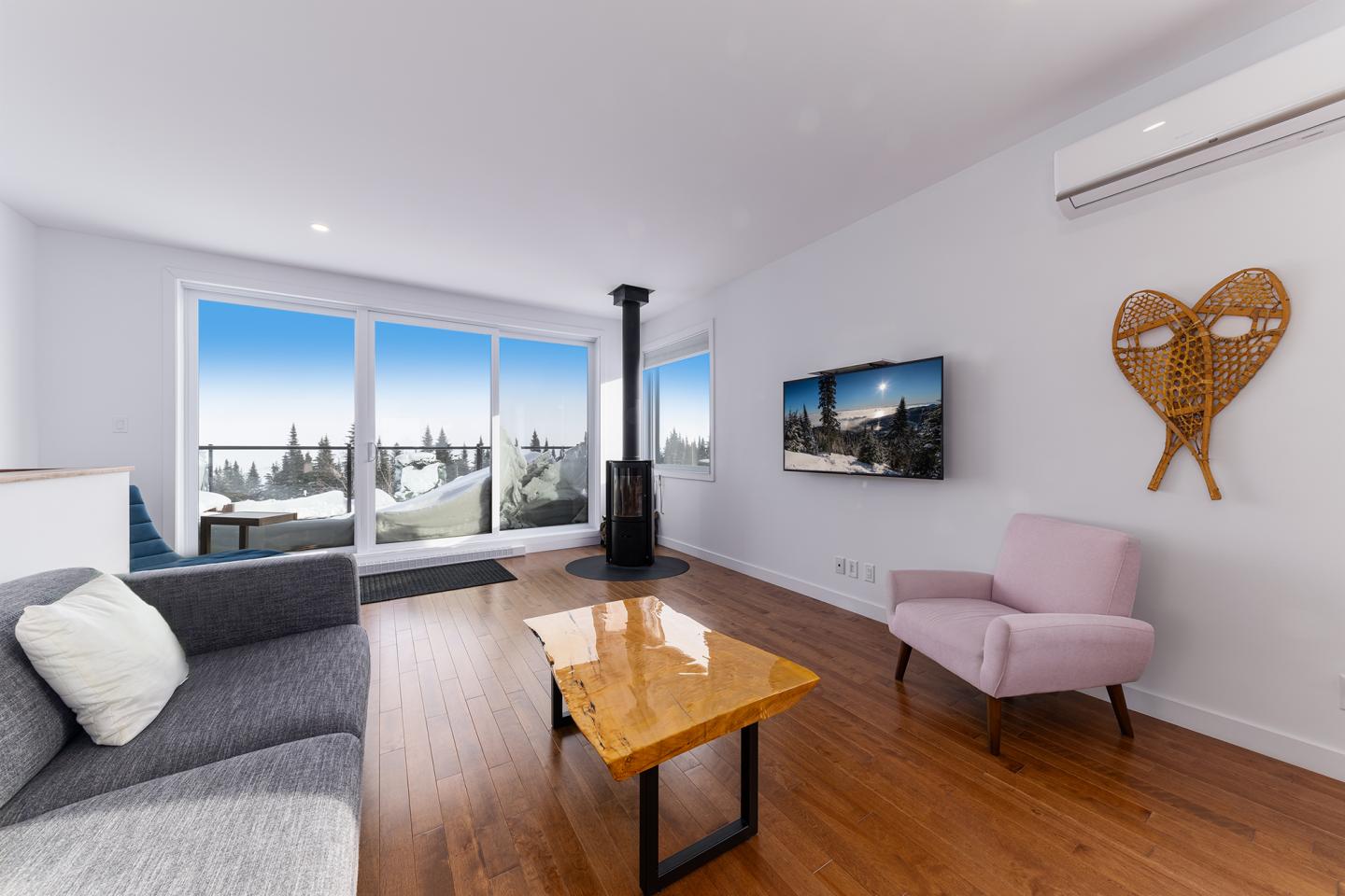 Salon moderne avec fenêtre sur paysage enneigé, poêle à bois, canapé gris, fauteuil rose.