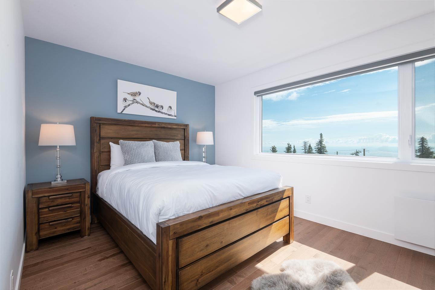 Chambre lumineuse avec lit en bois, mur bleu, et vue sur un paysage extérieur.