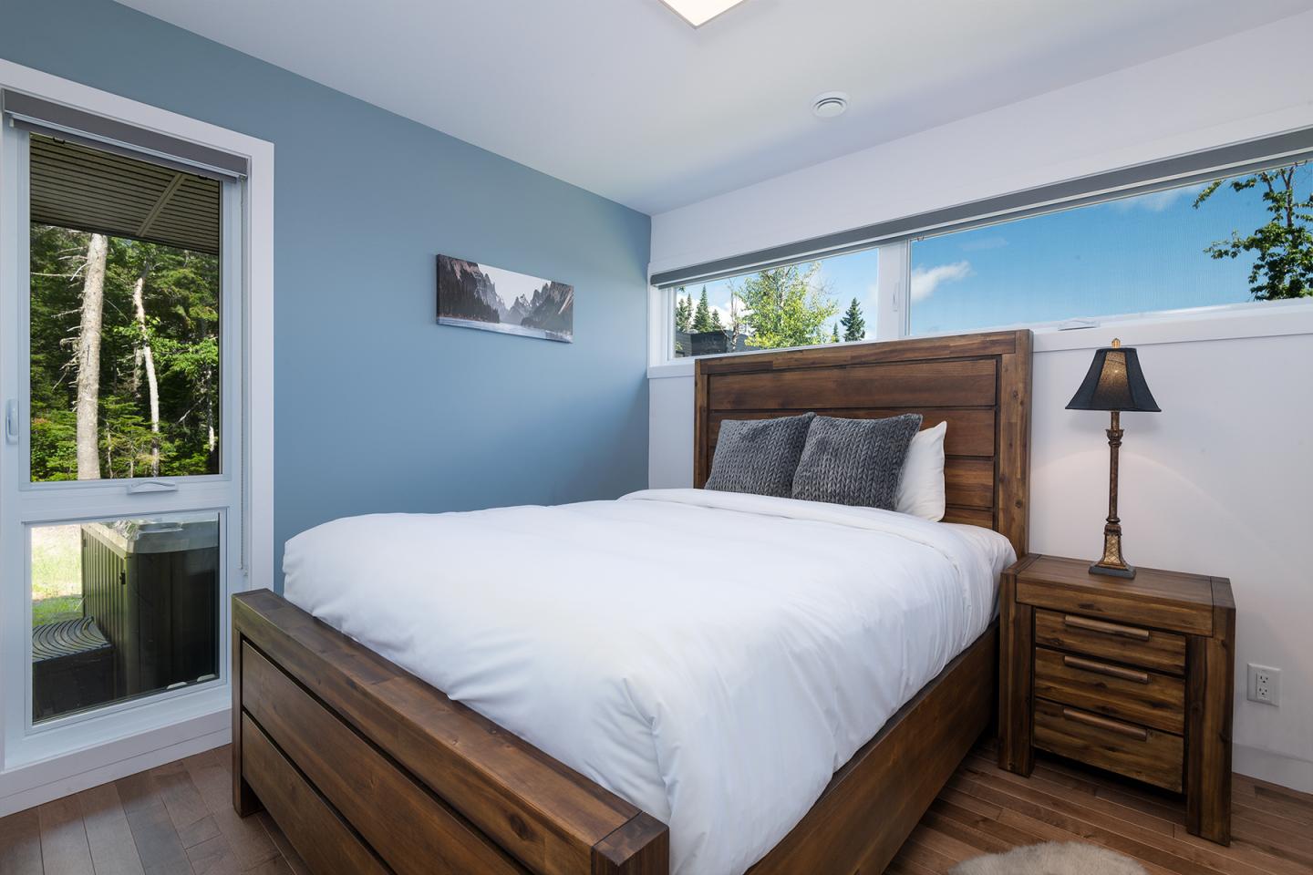Chambre lumineuse avec lit en bois, murs bleus, lampe et grande fenêtre.