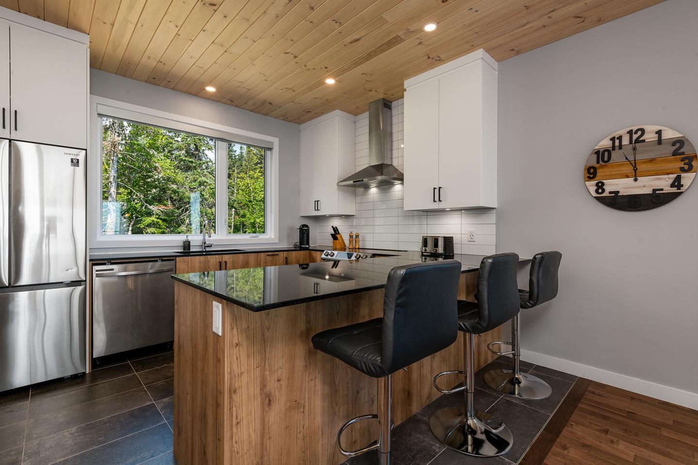 Cuisine moderne avec îlot central, chaises noires, et fenêtre donnant sur des arbres verts.