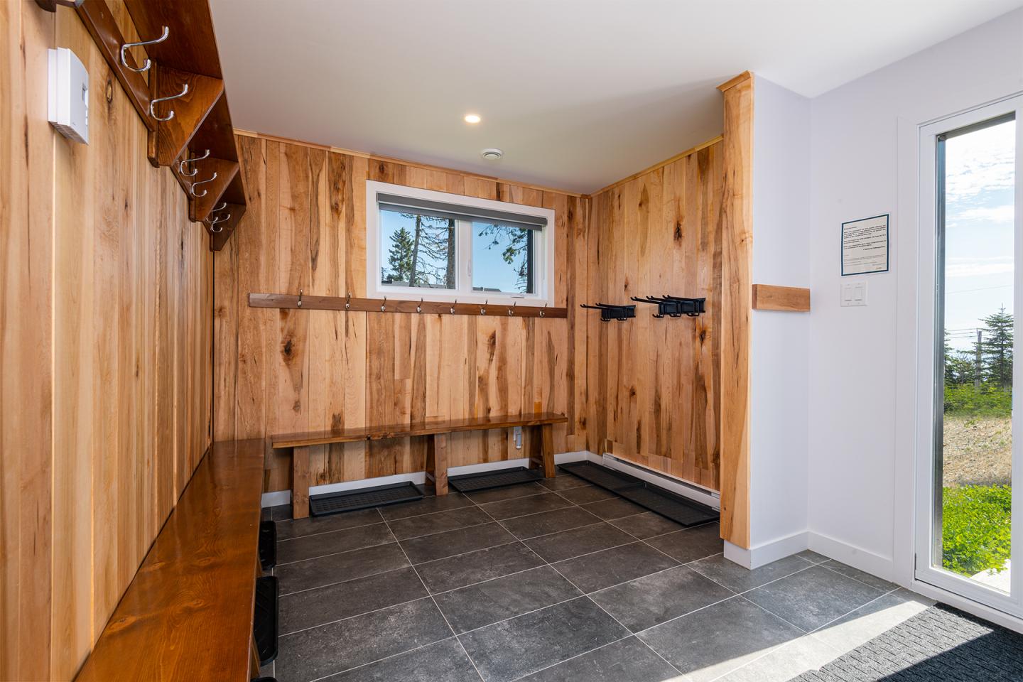 Vestiaire en bois clair avec porte-manteaux, fenêtre et sol en carrelage gris.