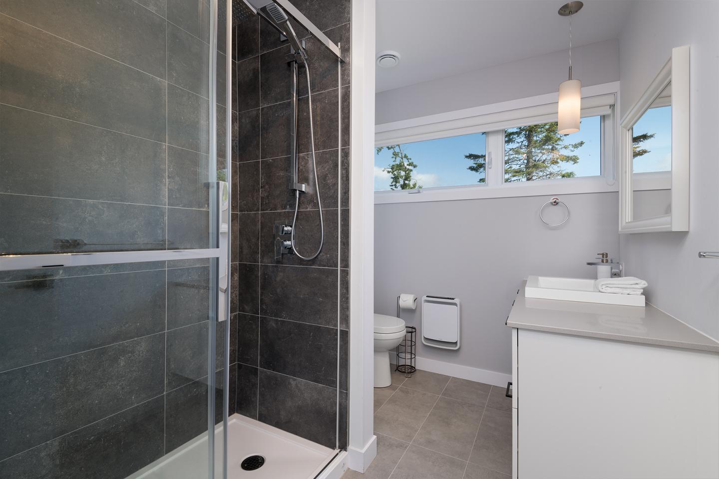 Salle de bain moderne, douche avec carrelage foncé, lavabo blanc, fenêtre lumineuse.