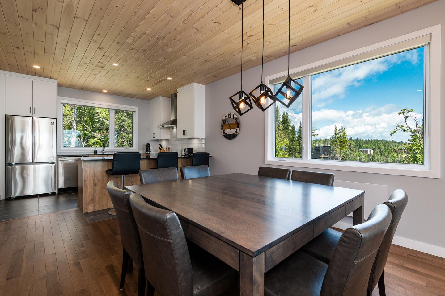 Salle à manger moderne avec table en bois, chaises en cuir, vue sur paysage boisé par la fenêtre.