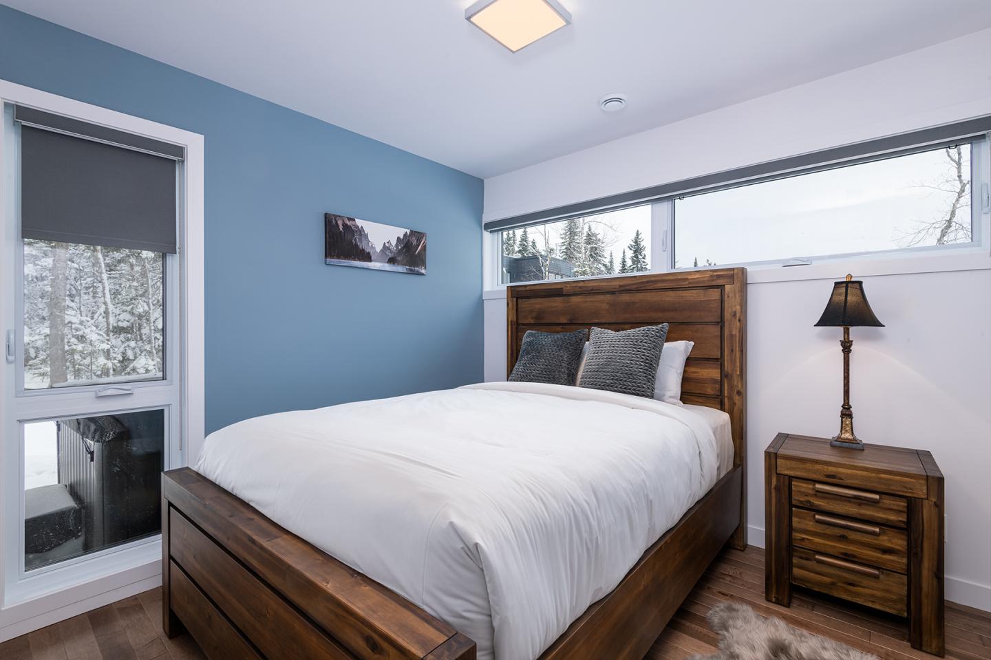 Chambre moderne avec lit en bois, murs bleus, lampe et fenêtre donnant sur l'extérieur.