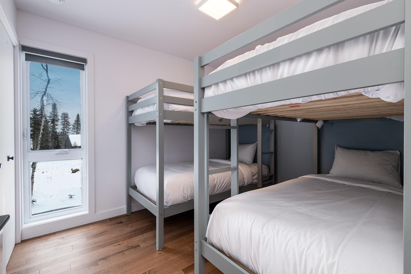 Chambre avec deux lits superposés gris, fenêtre vue sur neige et arbres.