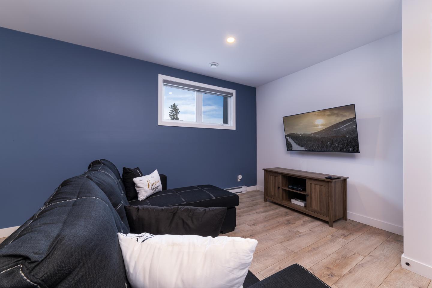Salon moderne avec mur bleu, canapé noir, télé et meuble bois. Fenêtre avec vue sur le ciel.