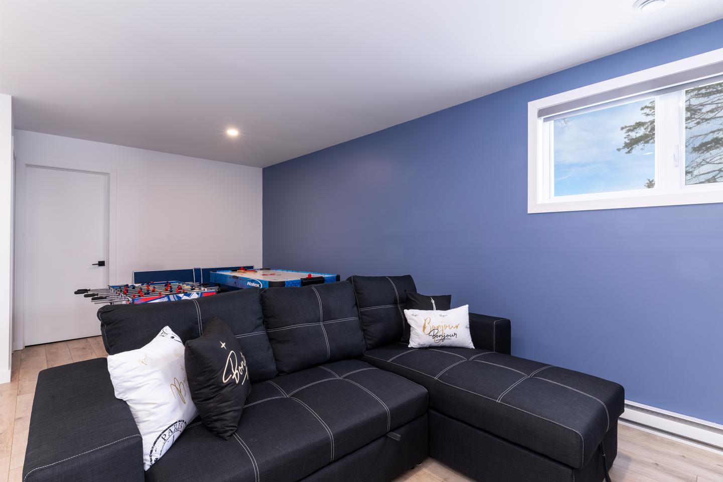 Salle avec canapé noir, table de baby-foot, mur bleu et fenêtre.