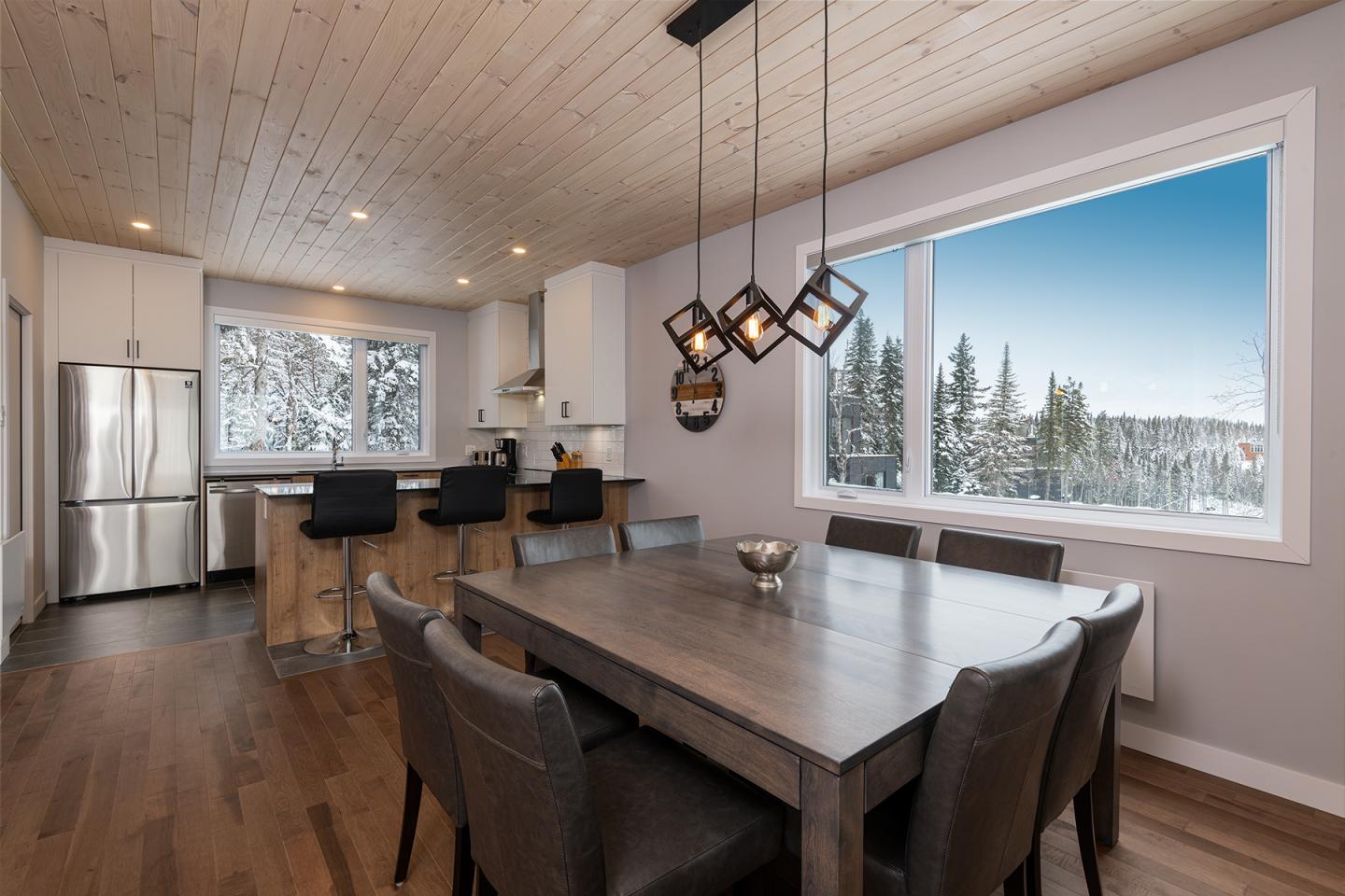Salle à manger moderne avec table en bois, chaises noires, cuisine ouverte. Fenêtre sur paysage enneigé.
