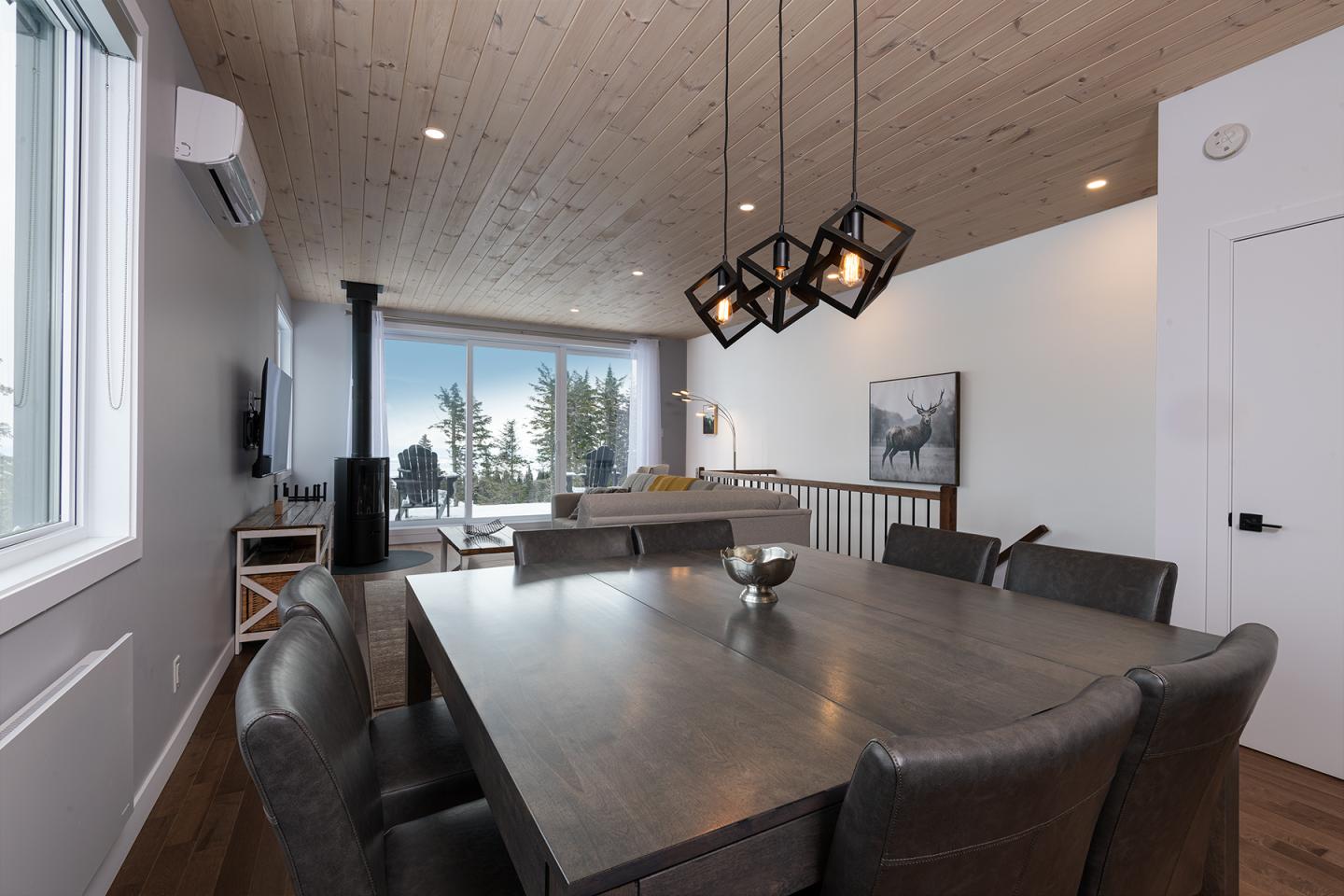 Salle à manger moderne avec table en bois foncé, luminaires suspendus, vue extérieure.