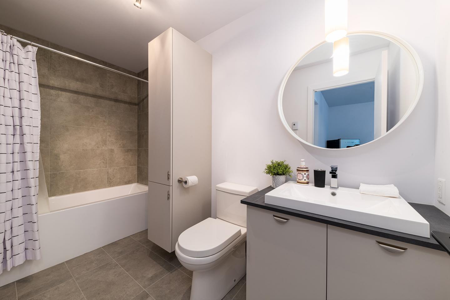 Salle de bain moderne avec lavabo, miroir rond, baignoire et rideau blanc.