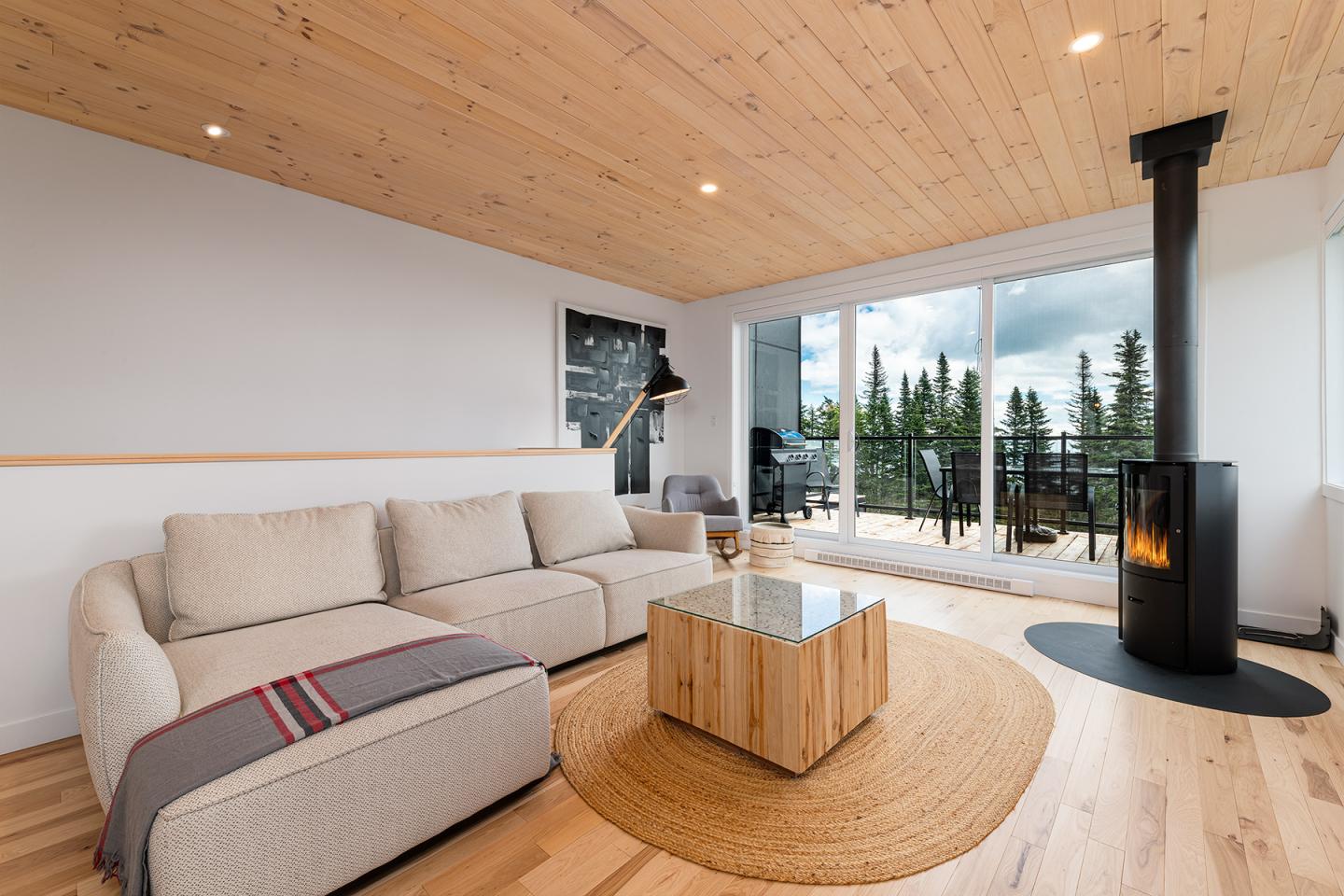 Salon moderne avec canapé beige, poêle à bois, grande baie vitrée, vue sur forêt.