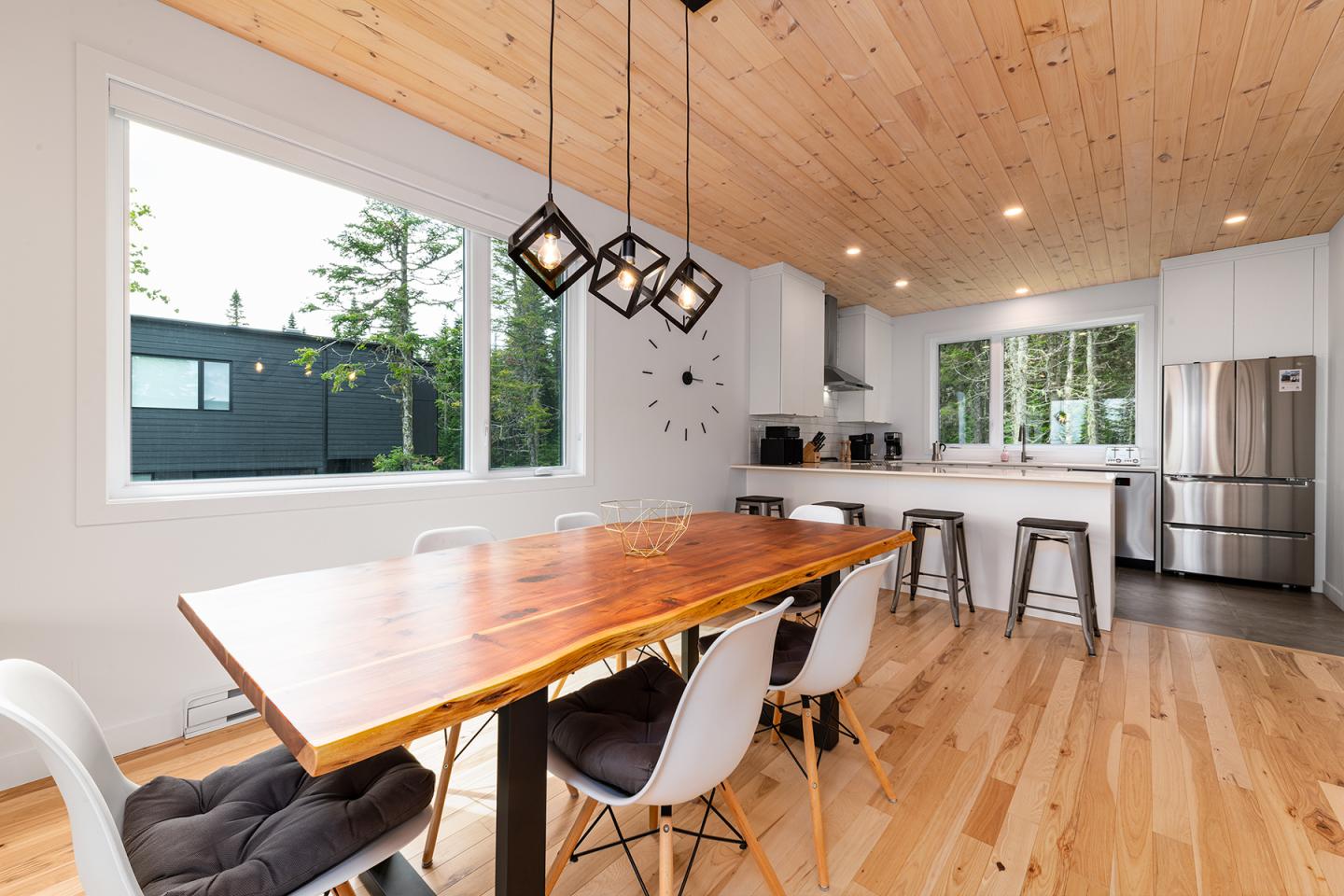 Salle à manger moderne avec table en bois, chaises blanches, cuisine ouverte et grandes fenêtres.