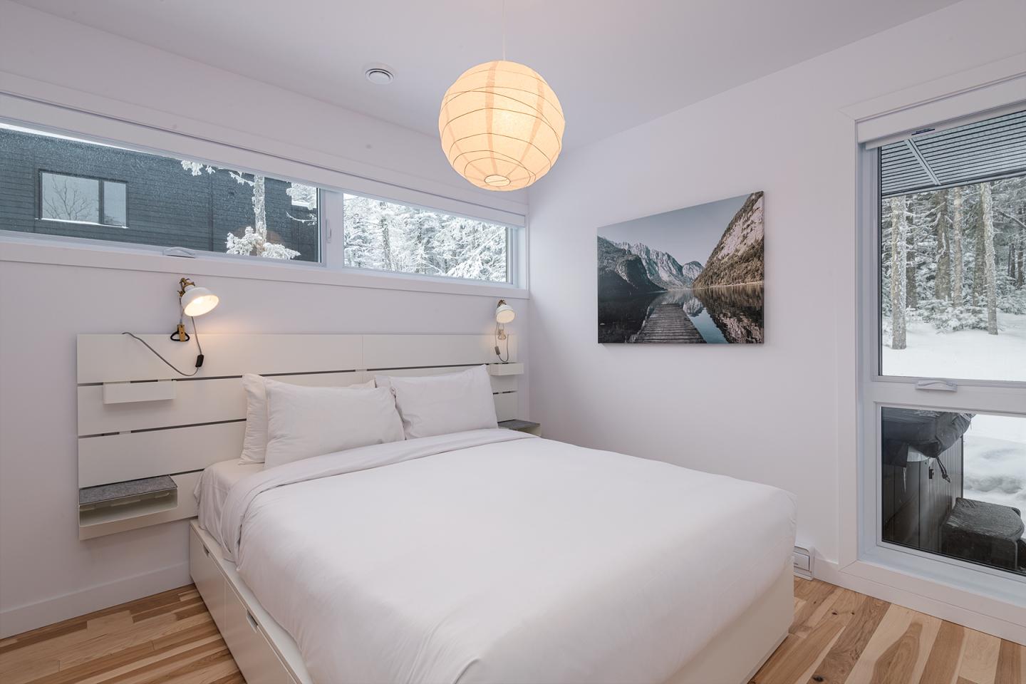 Chambre minimaliste blanche avec lit double et déco murale de montagne. Fenêtres enneigées.