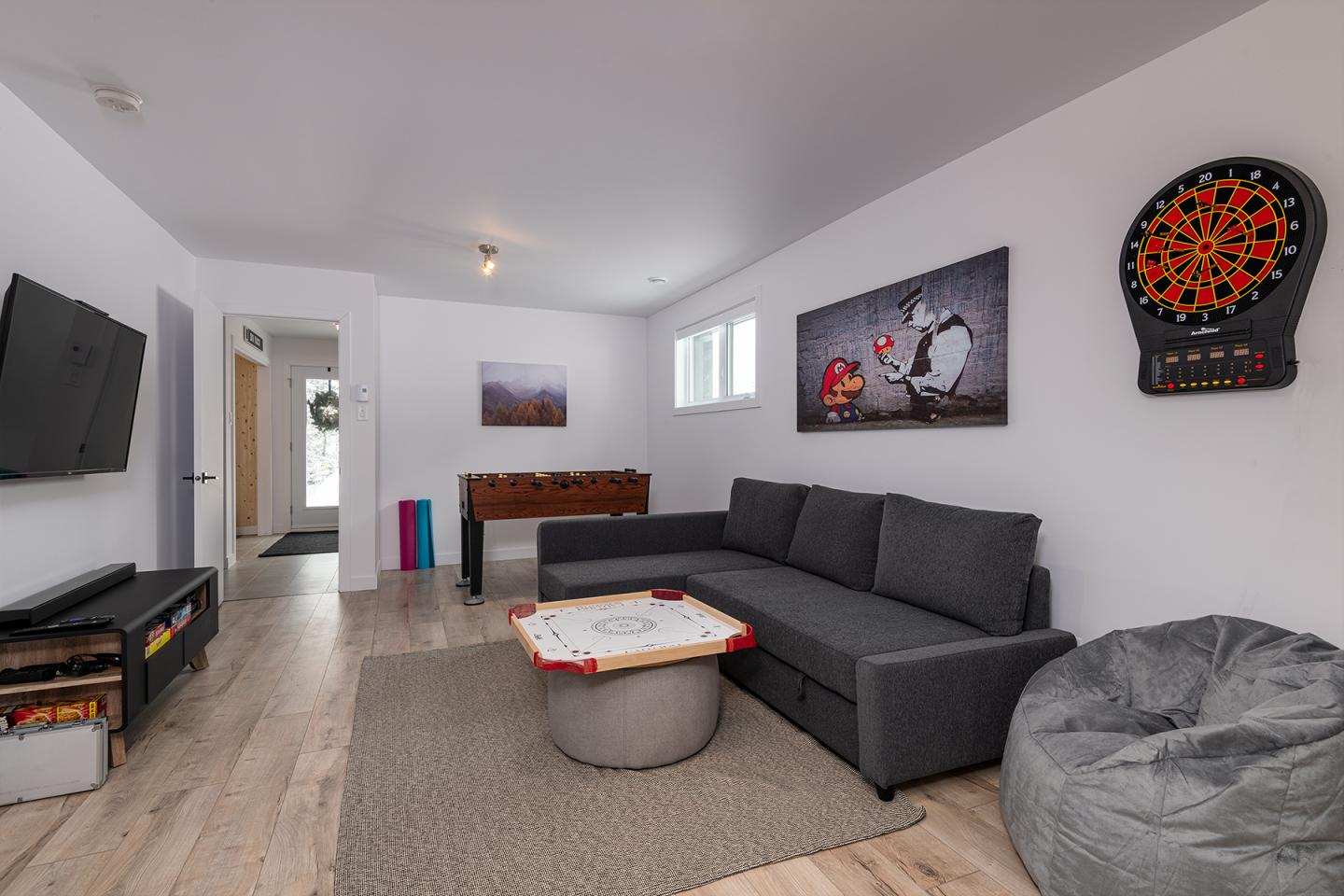 Salon moderne avec canapé gris, télé murale, baby-foot et cible de fléchettes.