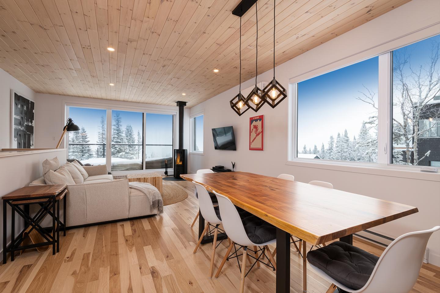 Salon moderne avec table en bois, canapé blanc et grandes fenêtres donnant sur un paysage enneigé.