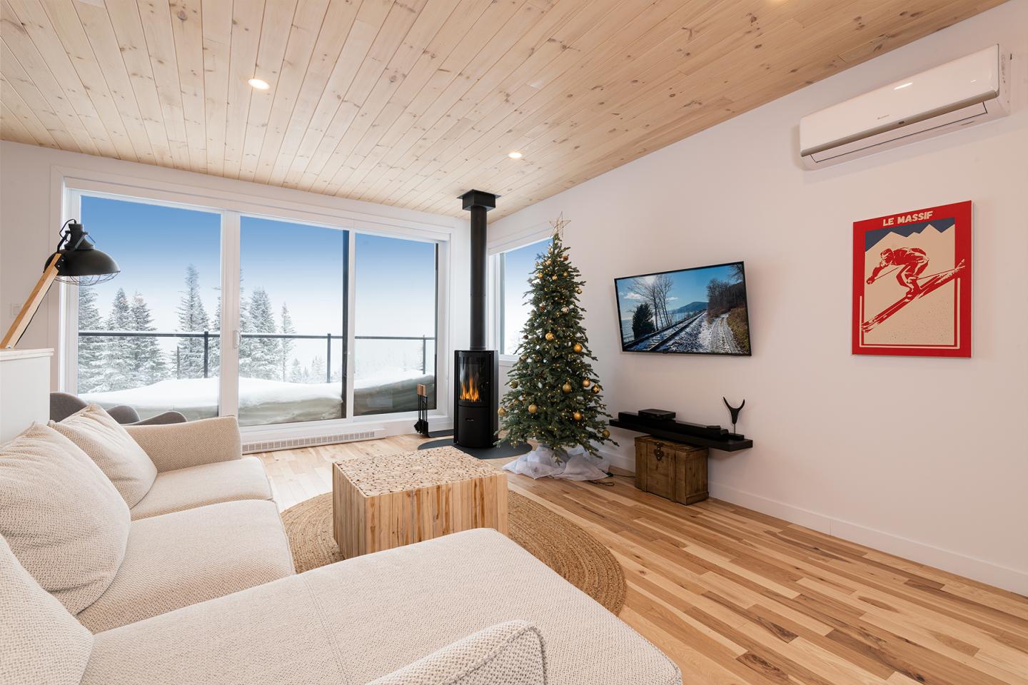 Salon cosy avec canapé, sapin de Noël, cheminée, et vue sur paysage enneigé par la fenêtre.