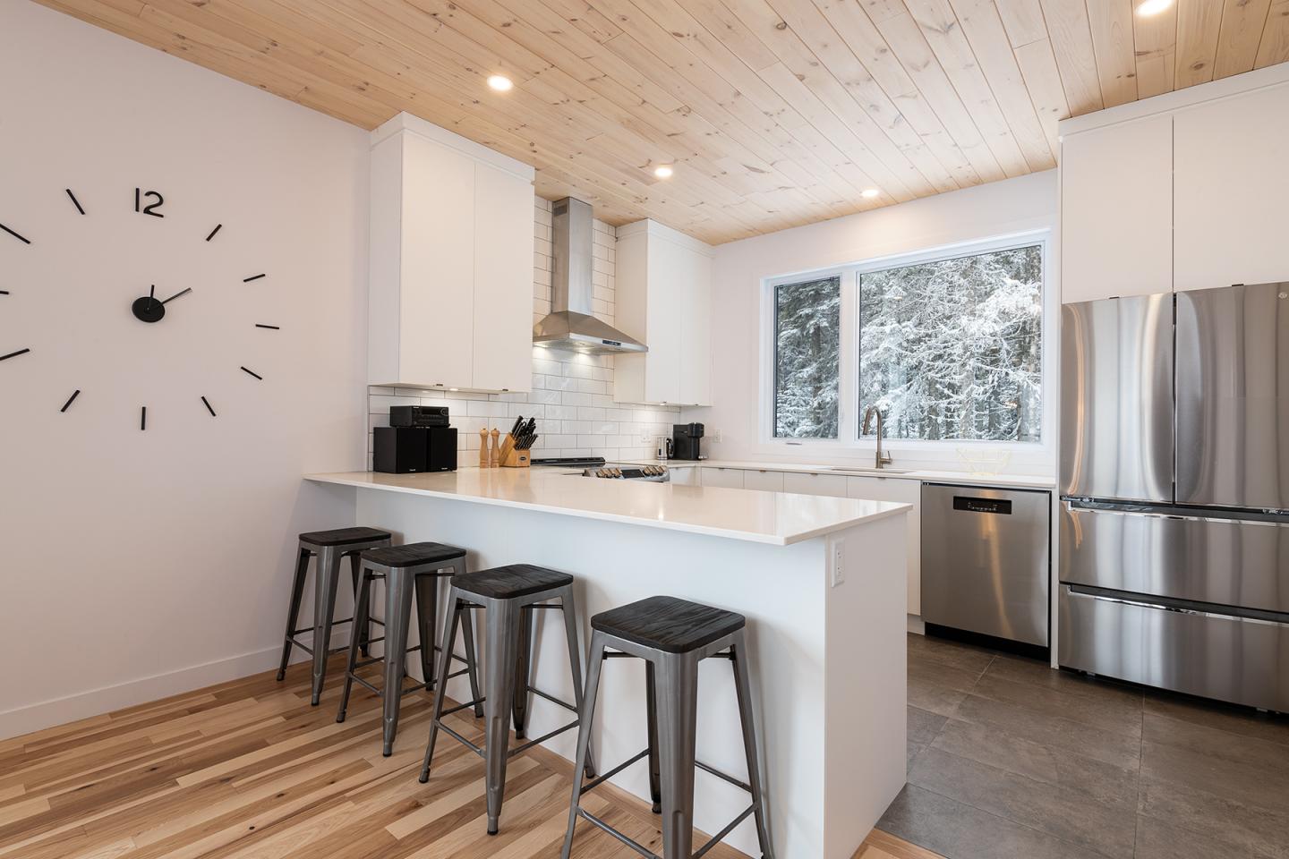 Cuisine moderne avec îlot, trois tabourets, four inox, et fenêtre avec vue sur arbres enneigés.