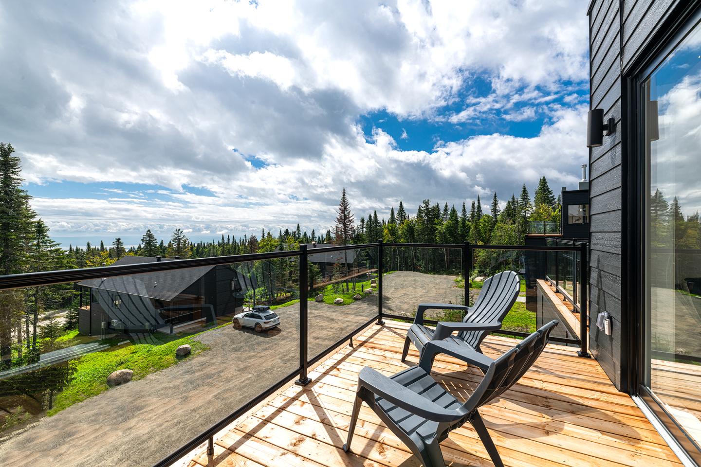 Terrasse en bois avec fauteuils, vue sur forêt et ciel partiellement nuageux.