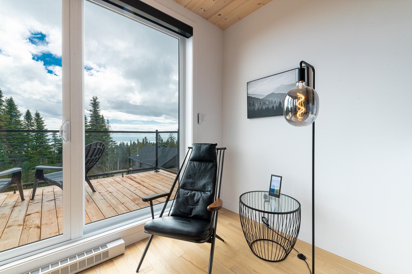 Fauteuil noir près d'une fenêtre avec vue sur terrasse boisée. Lampadaire moderne à droite.