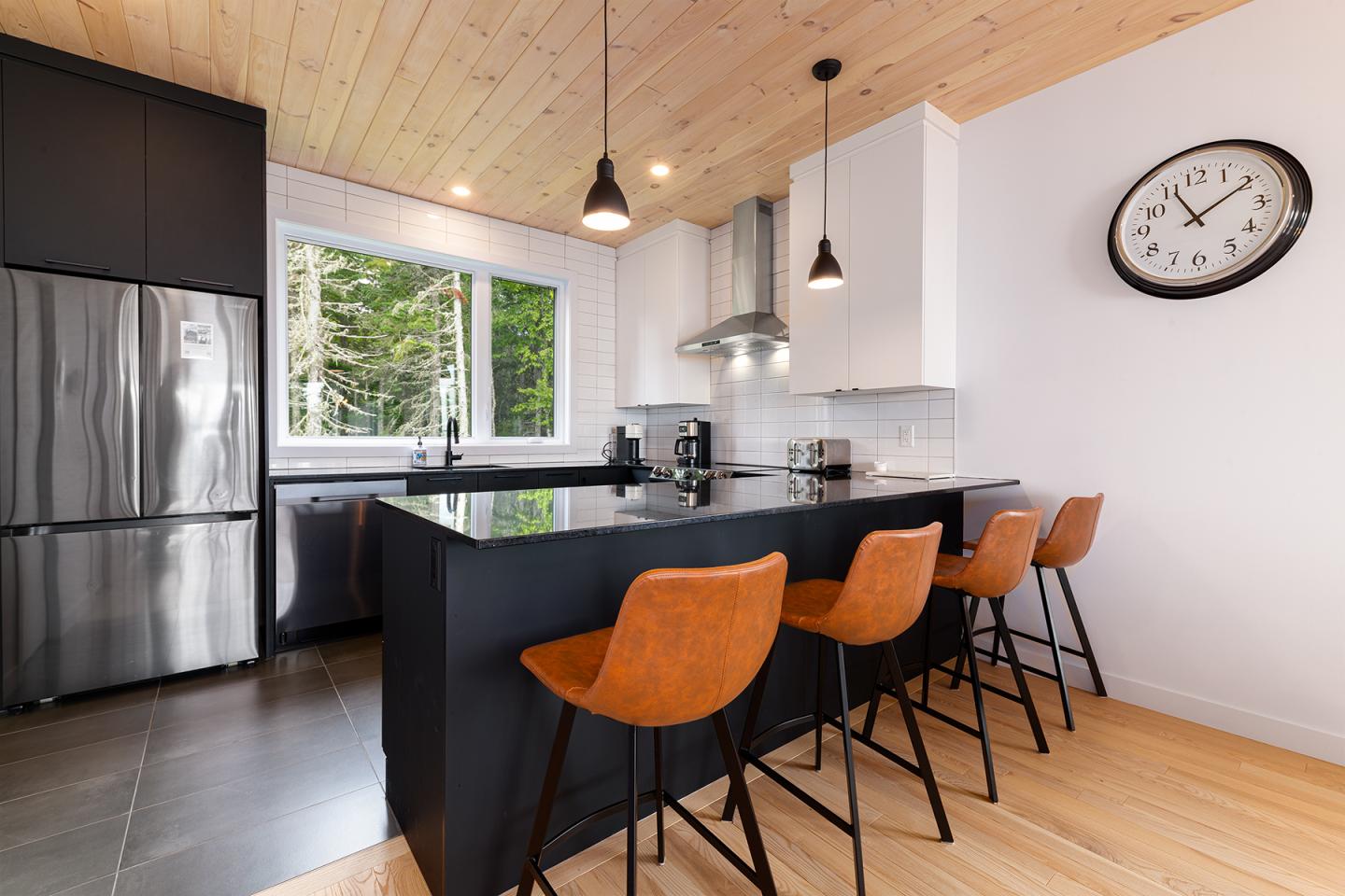 Cuisine moderne avec un îlot noir, tabourets orange et fenêtre avec vue sur des arbres.