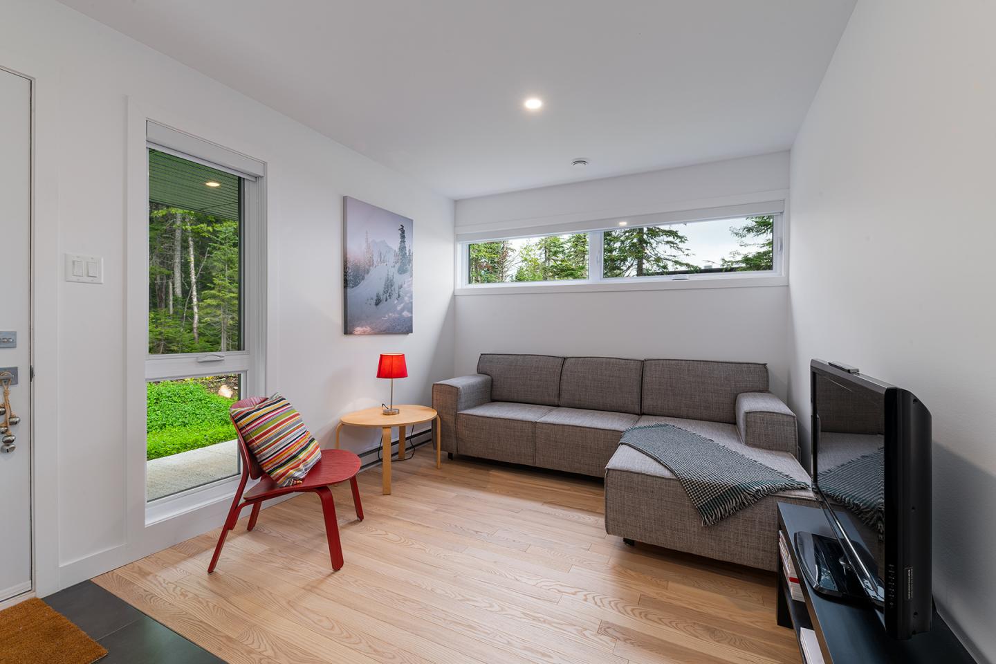 Salon moderne avec canapé gris, chaise rouge, et lampe rouge, vue sur jardin verdoyant.