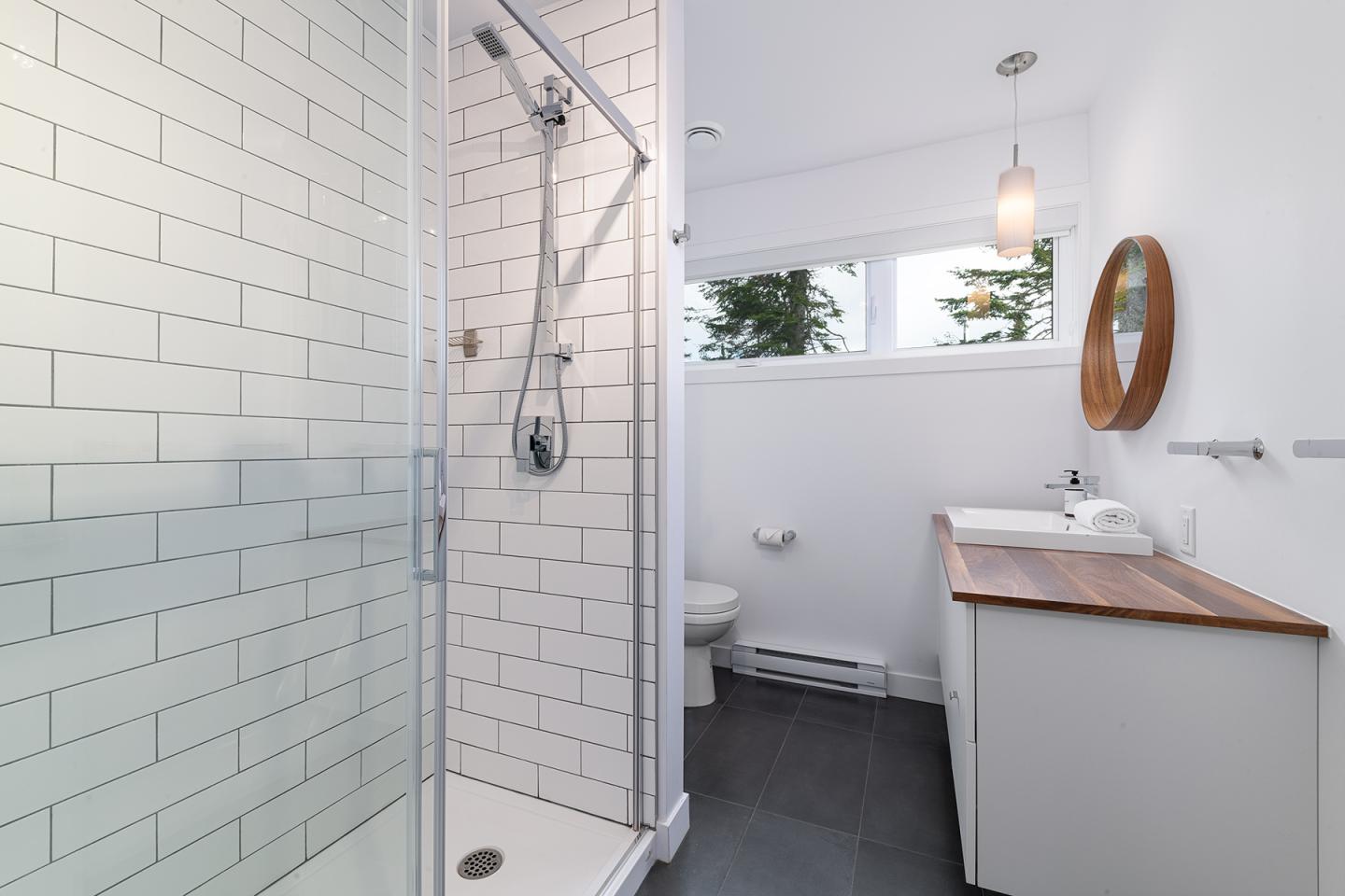 Salle de bain moderne, douche vitrée, lavabo blanc, miroir rond, murs carrelés.