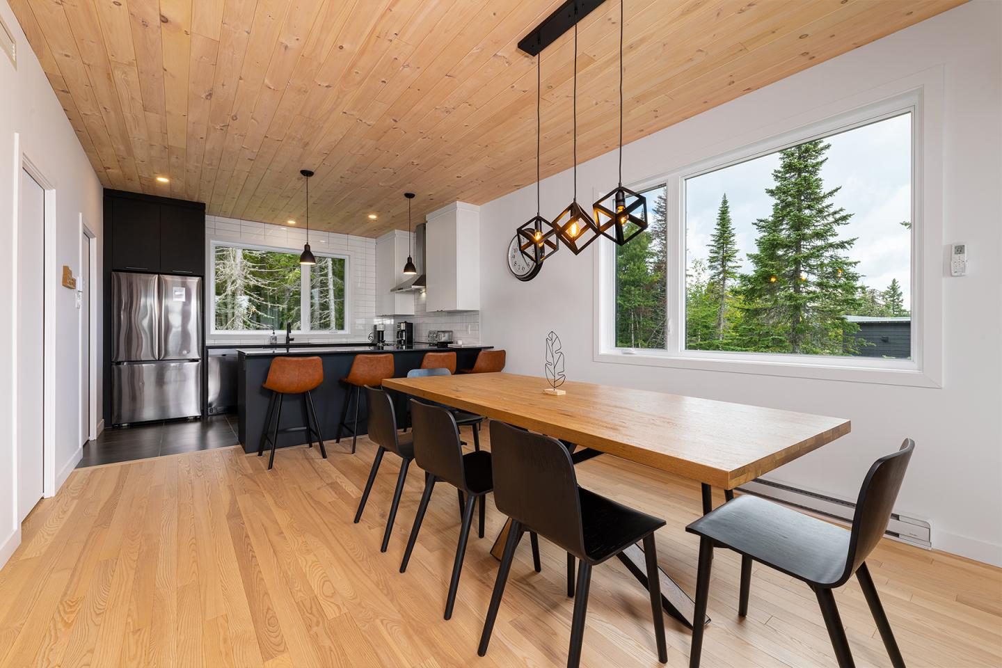 Salle à manger moderne avec grande table en bois, chaises noires et fenêtres donnant sur la forêt.