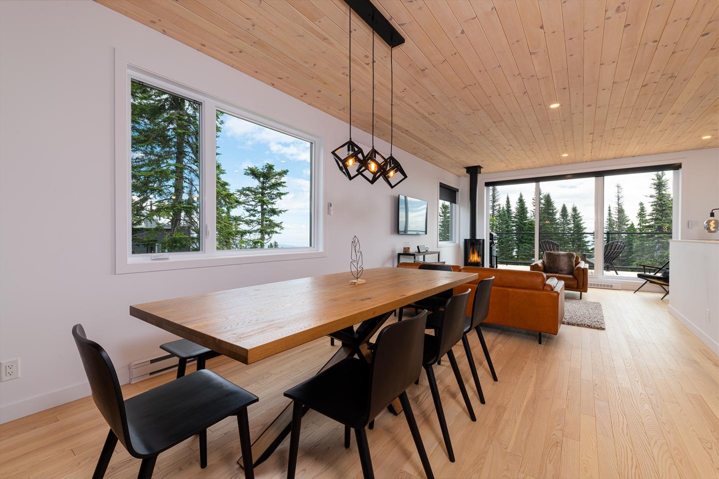 Salle à manger moderne avec table en bois, chaises noires et vue sur forêt par grandes fenêtres.