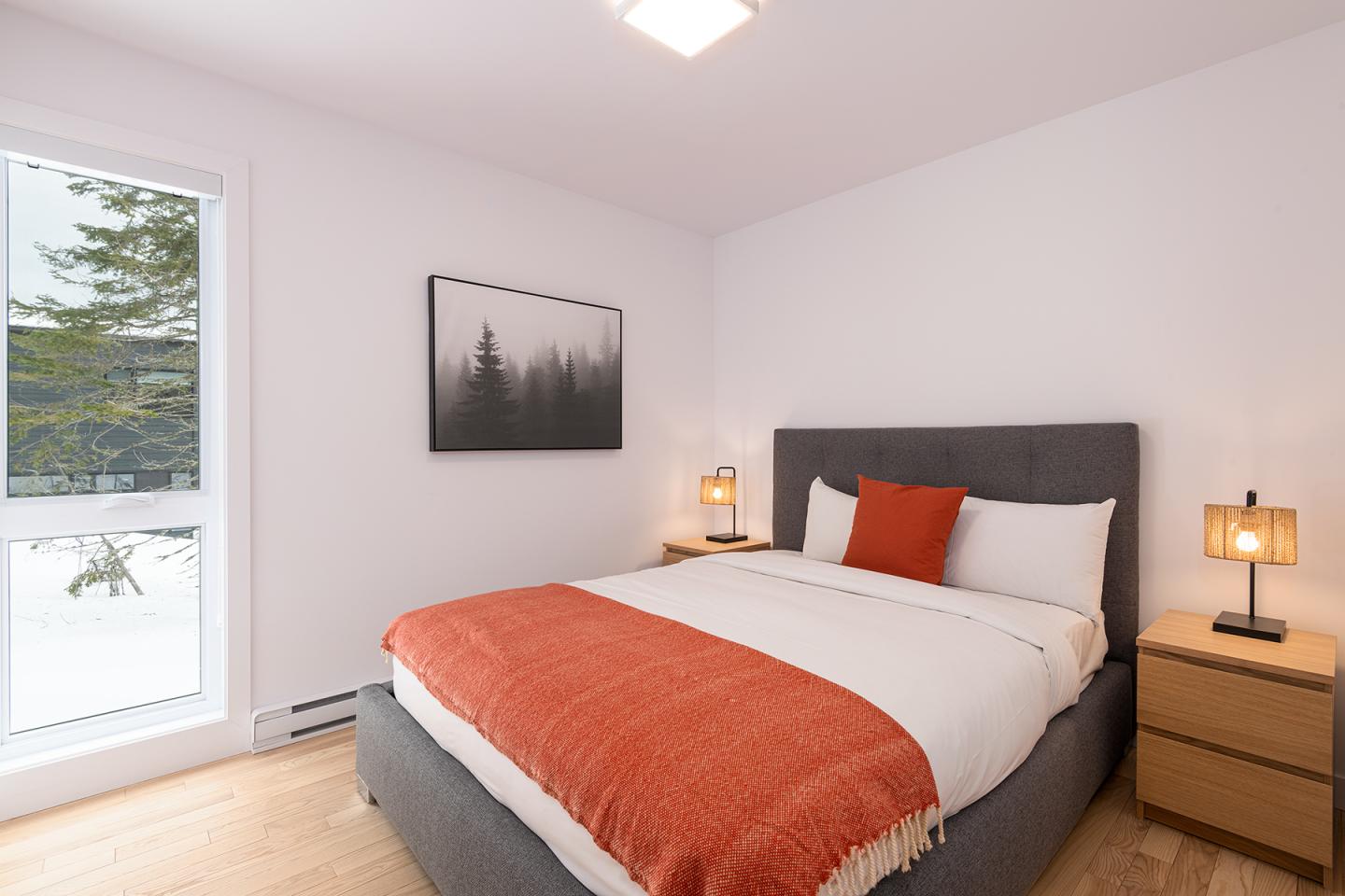 Chambre moderne avec lit gris et couverture orange. Tableau de forêt et lampes sur chevet.
