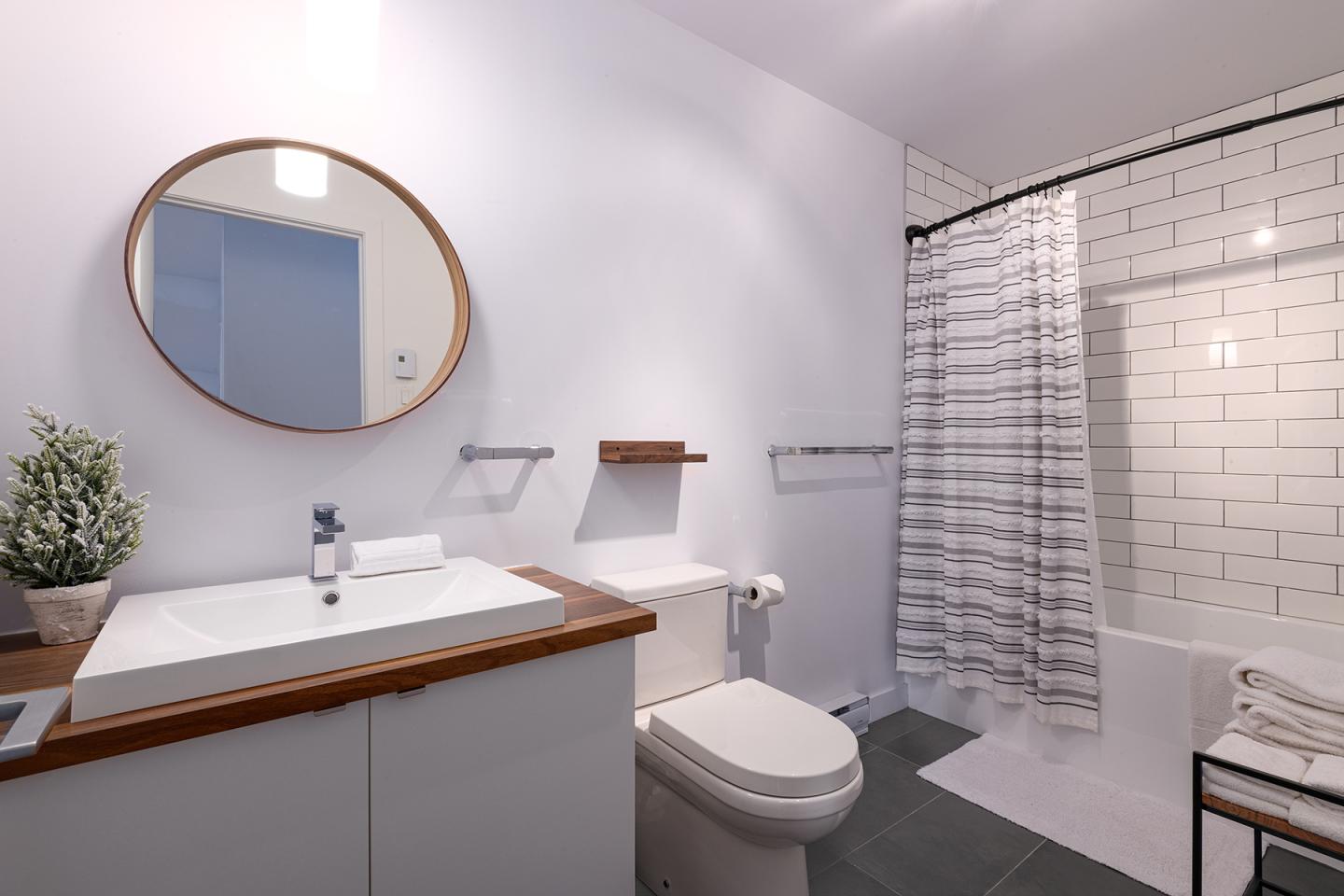 Salle de bain moderne avec lavabo, miroir rond, douche et carrelage blanc.