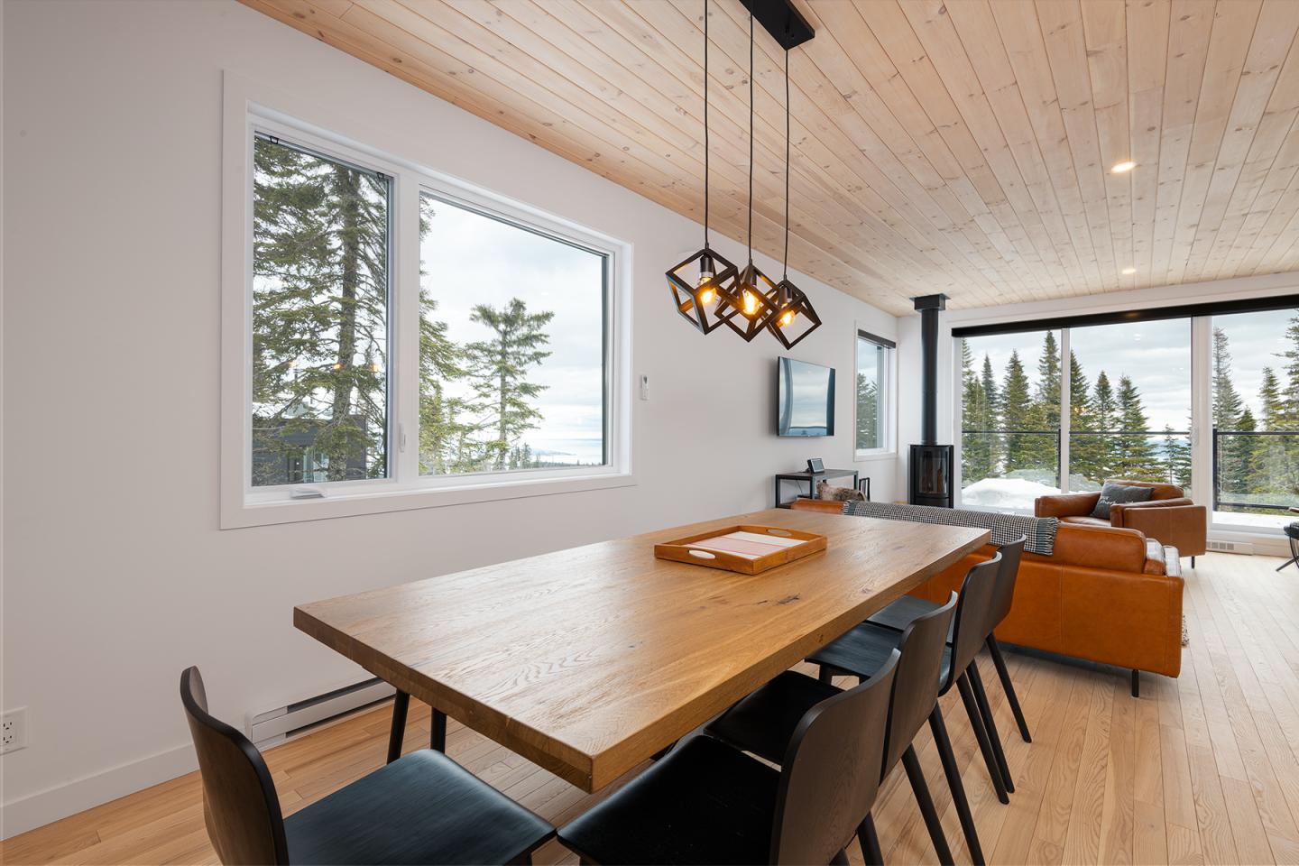 Salle à manger moderne avec table en bois, chaises noires, grandes fenêtres sur forêt.