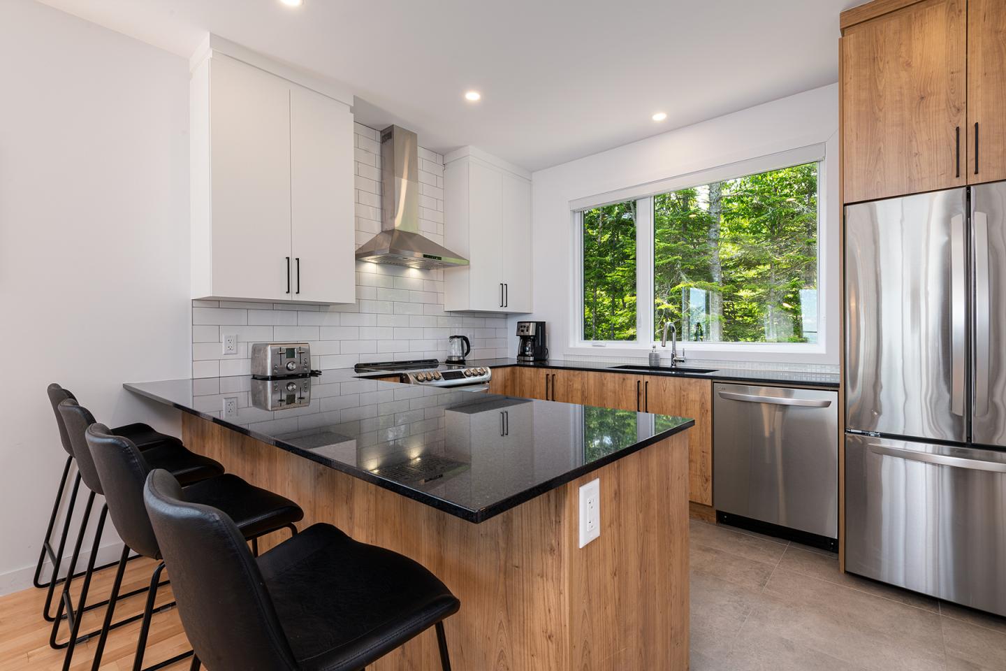 Cuisine moderne avec comptoir noir, tabourets, et électroménagers inox, grande fenêtre.