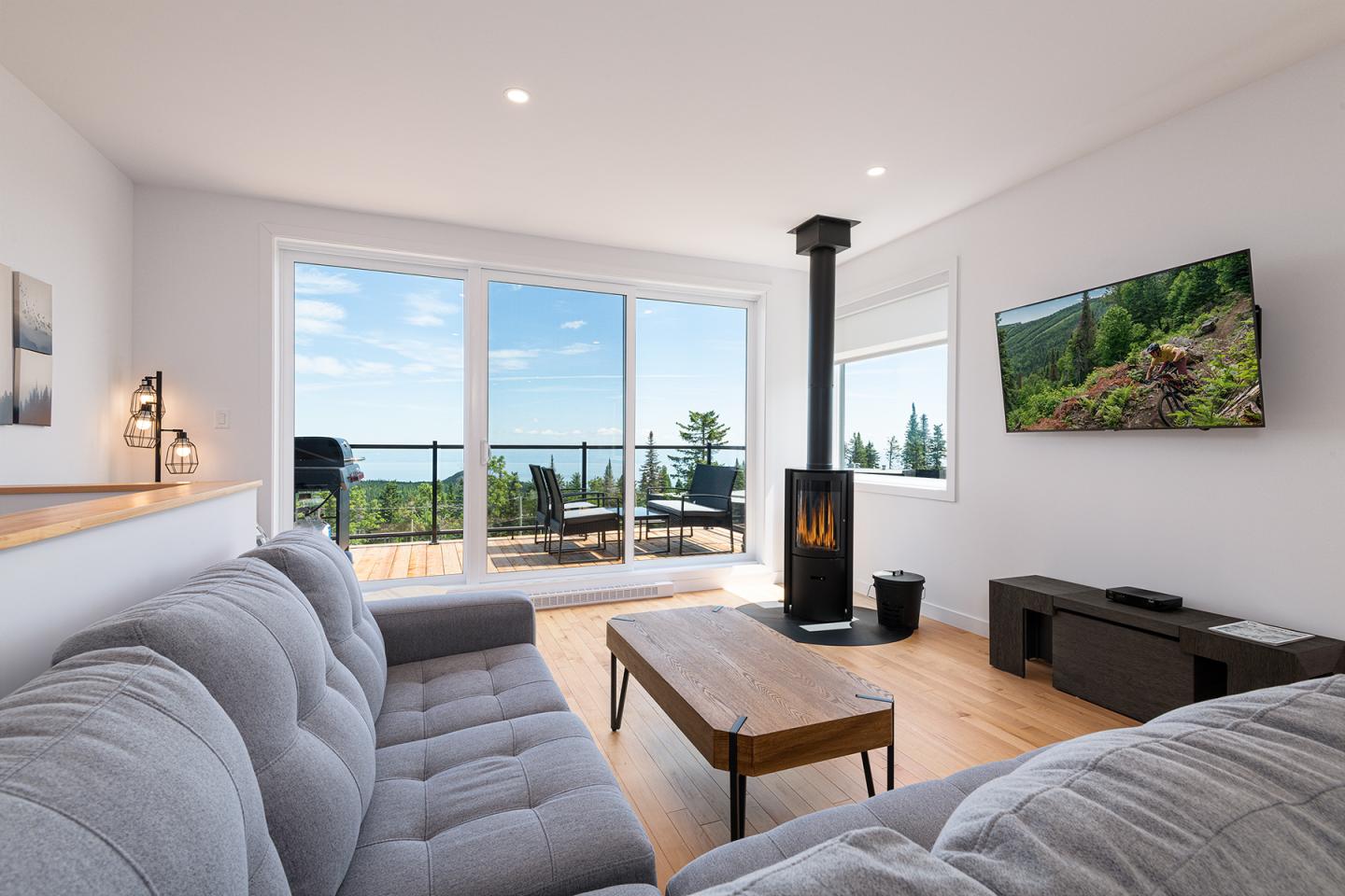 Salon moderne avec canapé gris et poêle à bois, vue sur terrasse et forêt.