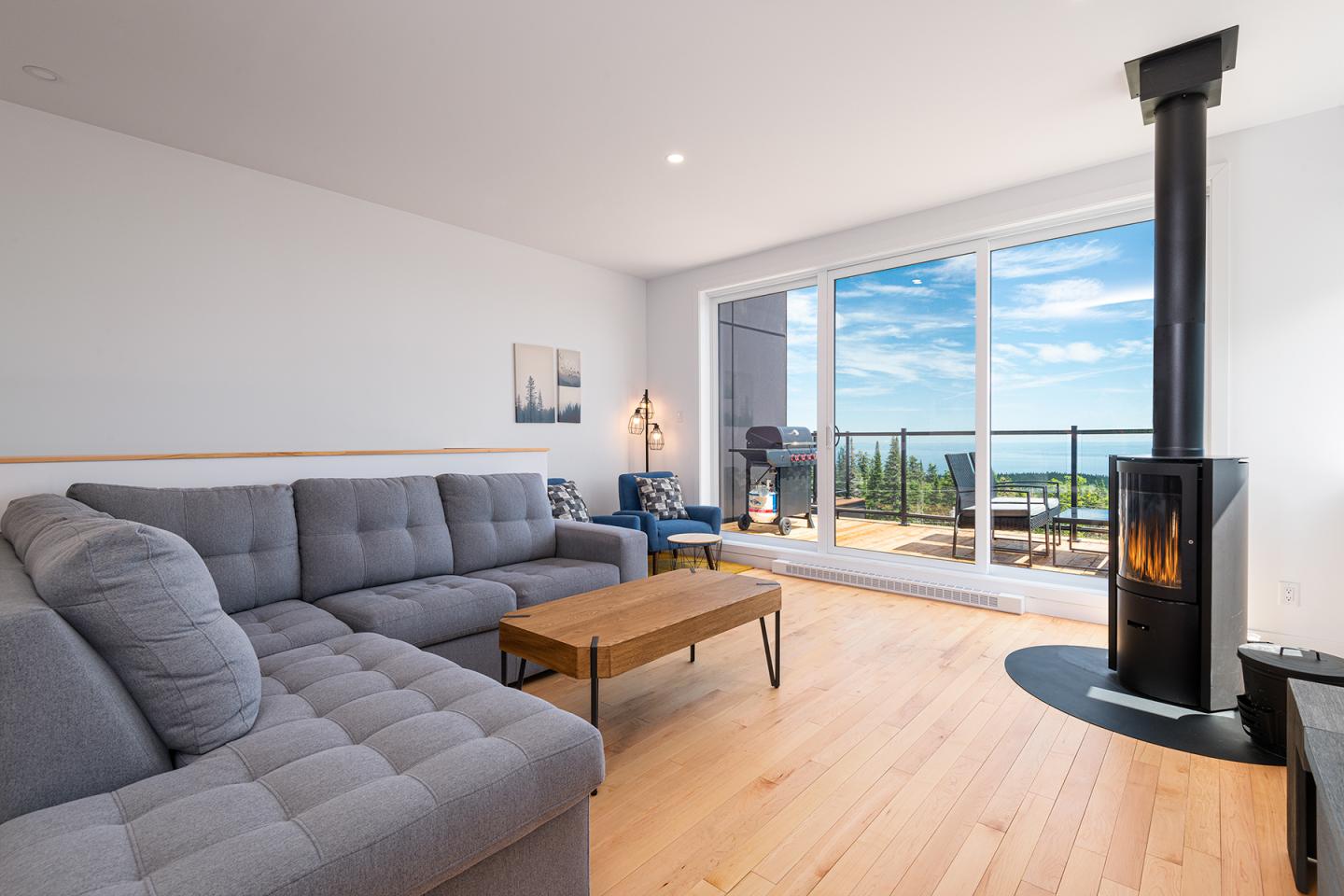 Salon moderne avec canapé gris, poêle, et vue sur mer à travers une baie vitrée.