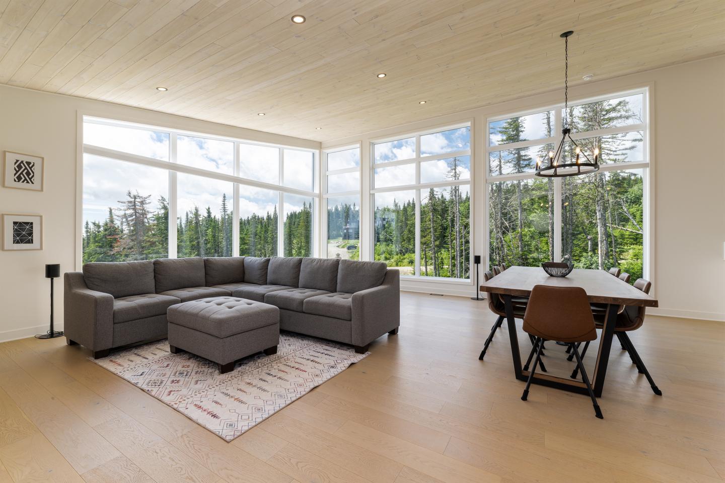 Salon moderne avec canapé gris, table à manger et grandes fenêtres donnant sur la forêt.