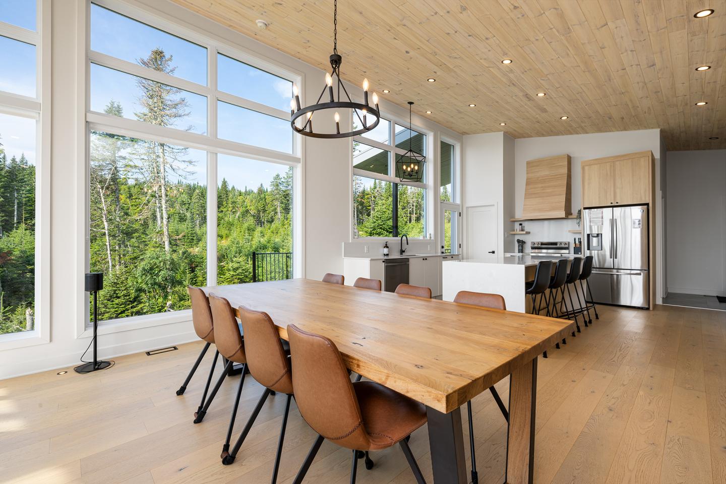 Salle à manger moderne avec grande table en bois, chaises marron, et cuisine ouverte lumineuse.