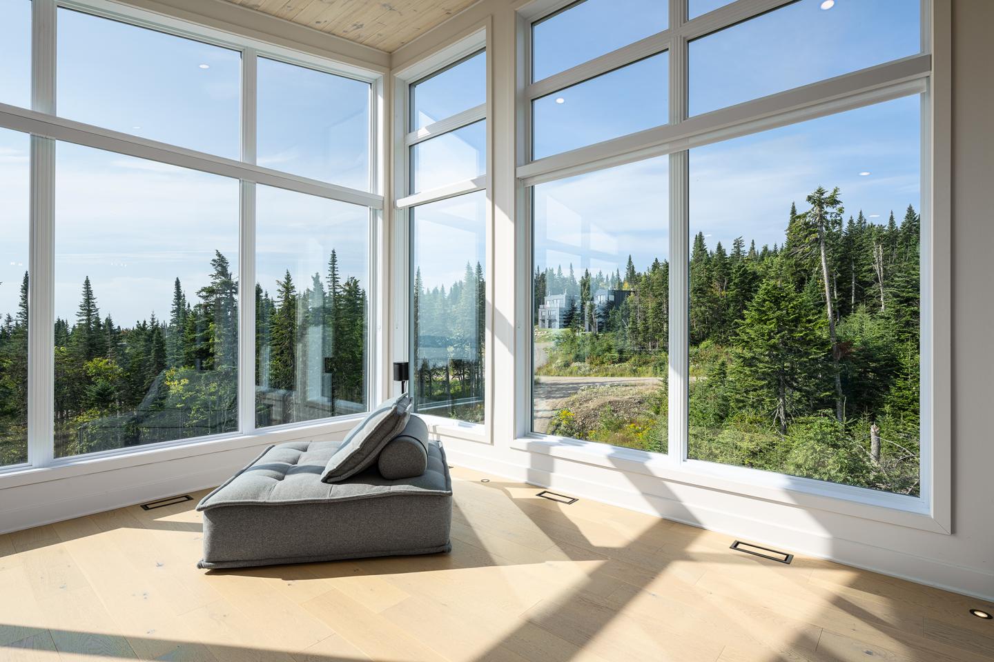 Salon moderne avec canapé gris, grandes fenêtres, vue sur la forêt.