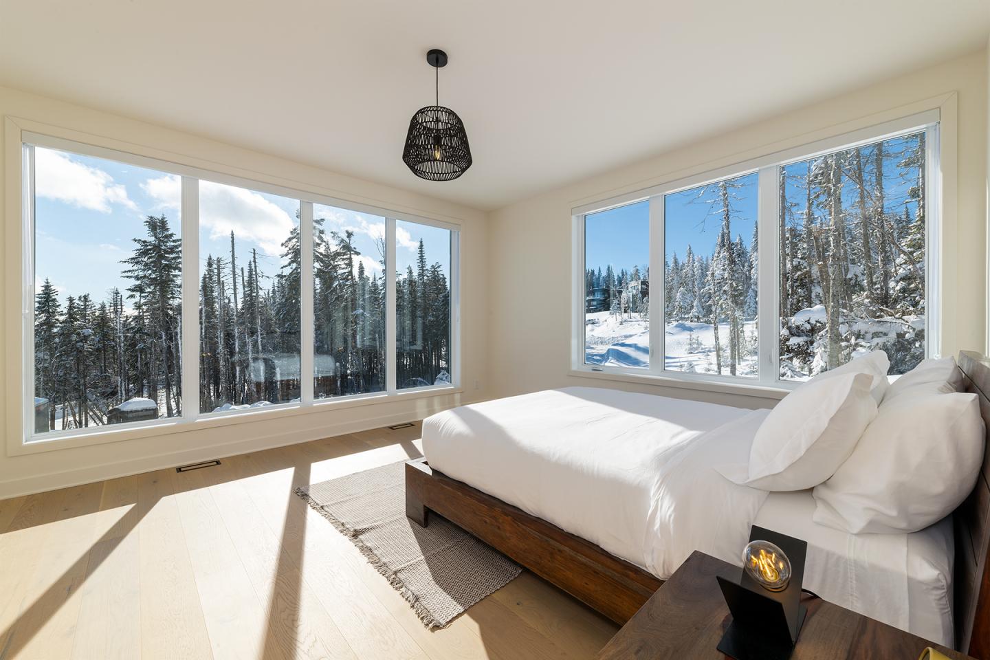 Chambre lumineuse avec lit blanc, grandes fenêtres donnant sur paysage enneigé.