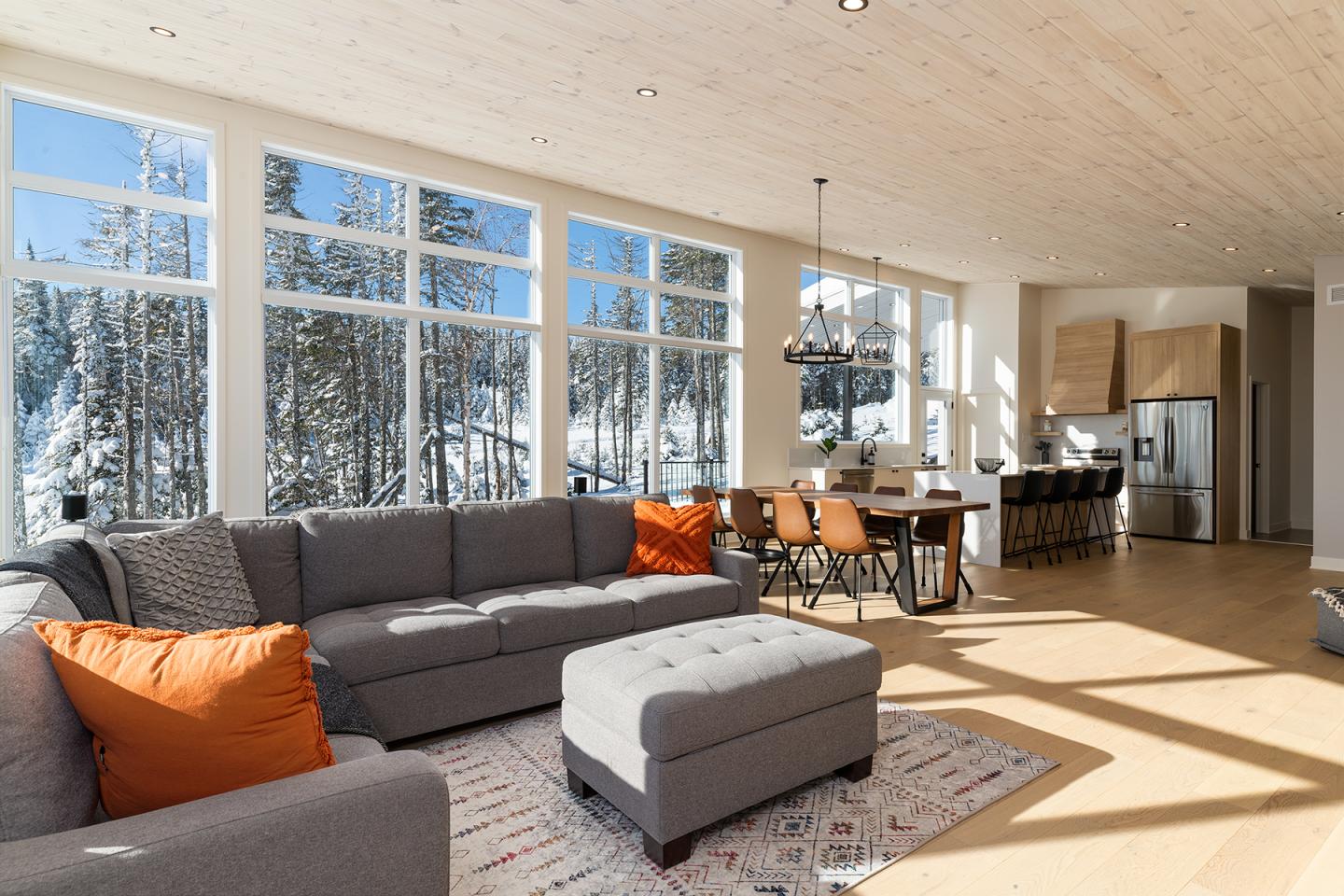 Salon moderne avec canapé gris, coussins orange, et grandes fenêtres donnant sur forêt enneigée.