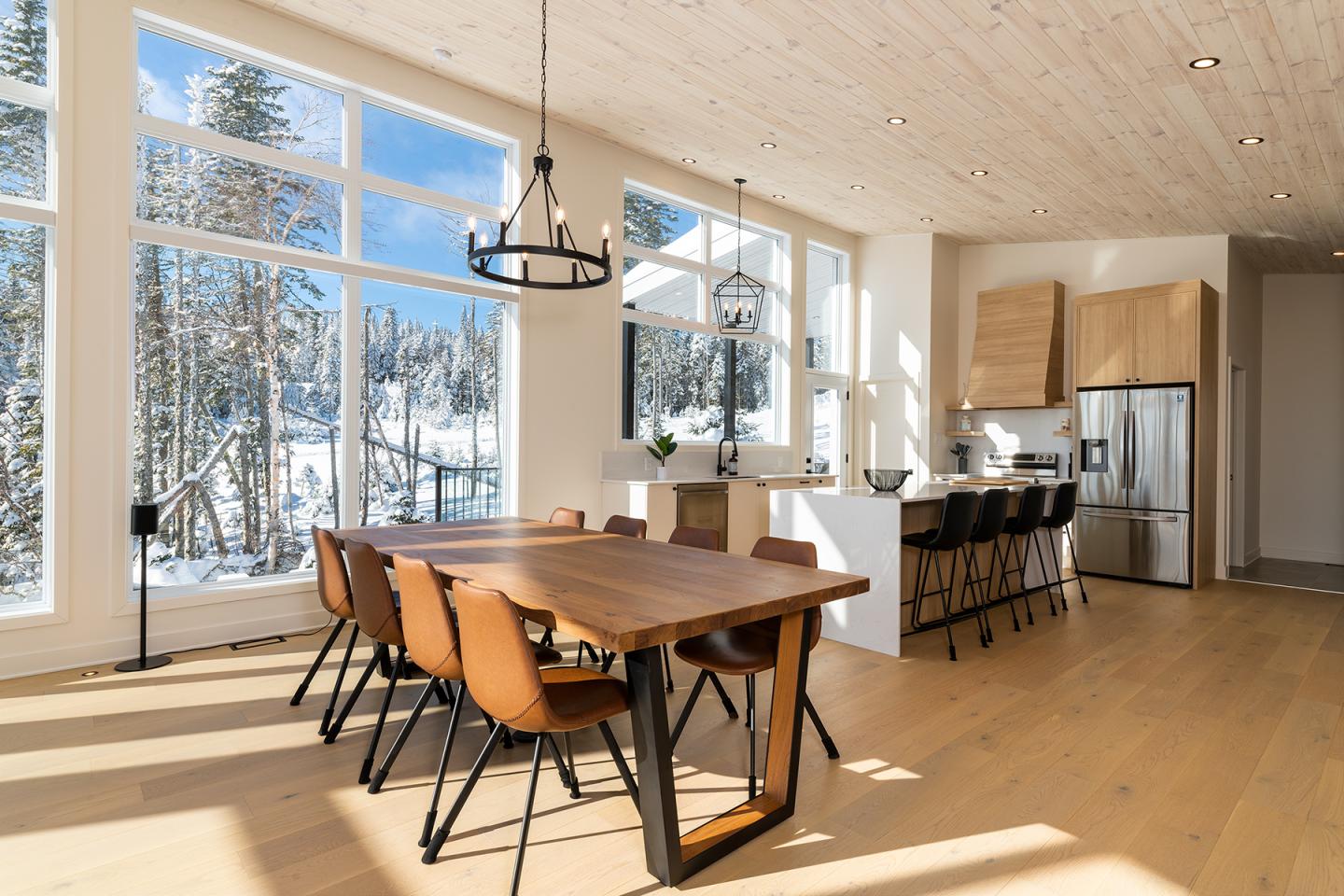 Salle à manger avec grande table en bois, cuisine à l'arrière, vue sur la neige à l'extérieur.